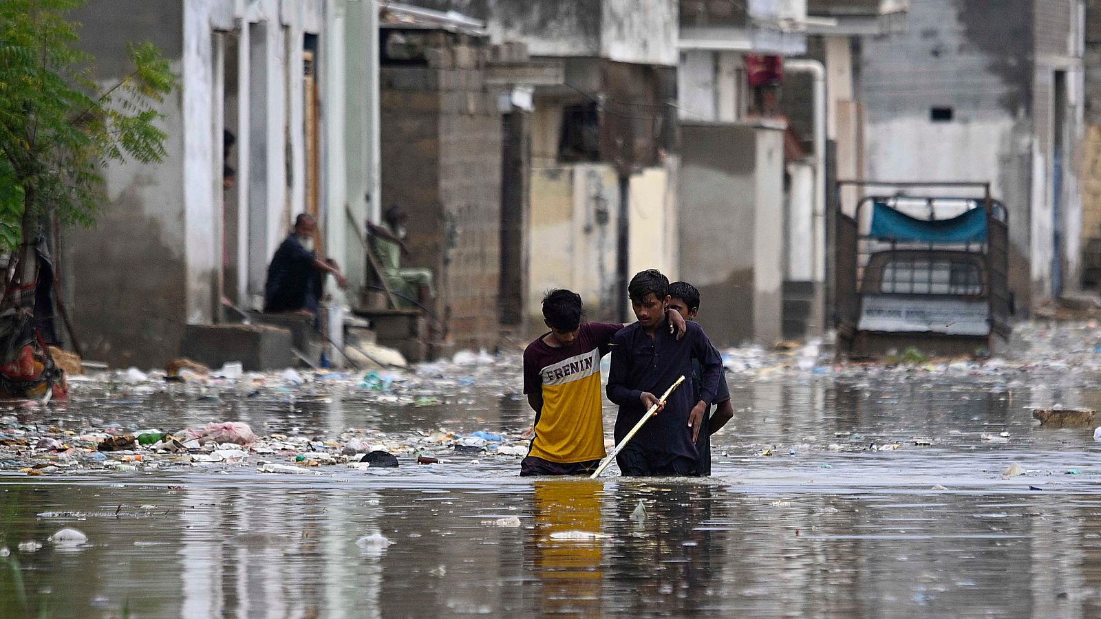 Niños caminando por calle inundada, agua a la cintura, ropa oscura y amarilla, casas deterioradas al fondo.