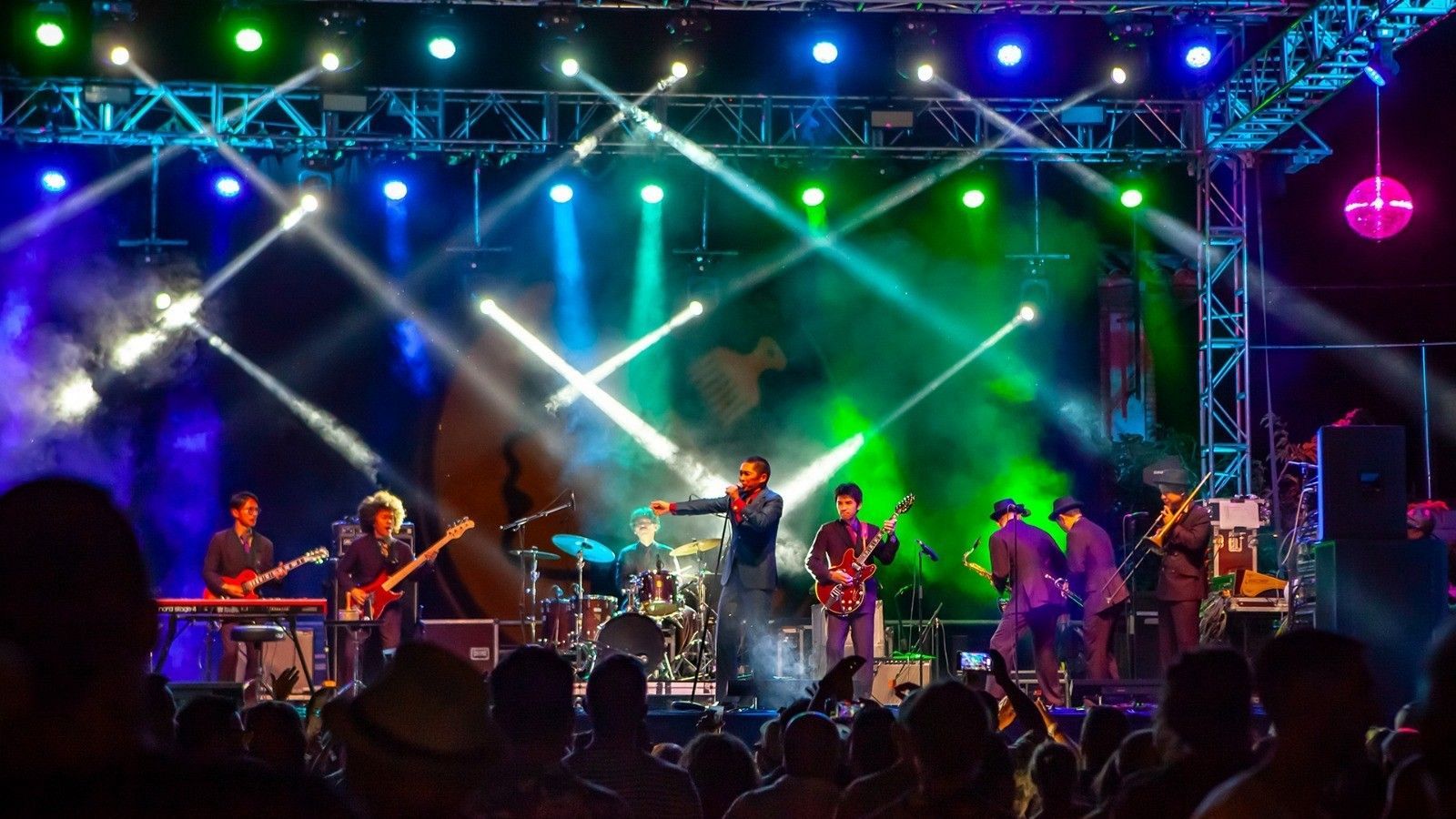 Actuación musical en vivo con varios músicos, luces de colores y público.  Se aprecia un cantante con traje azul y una sección de viento.