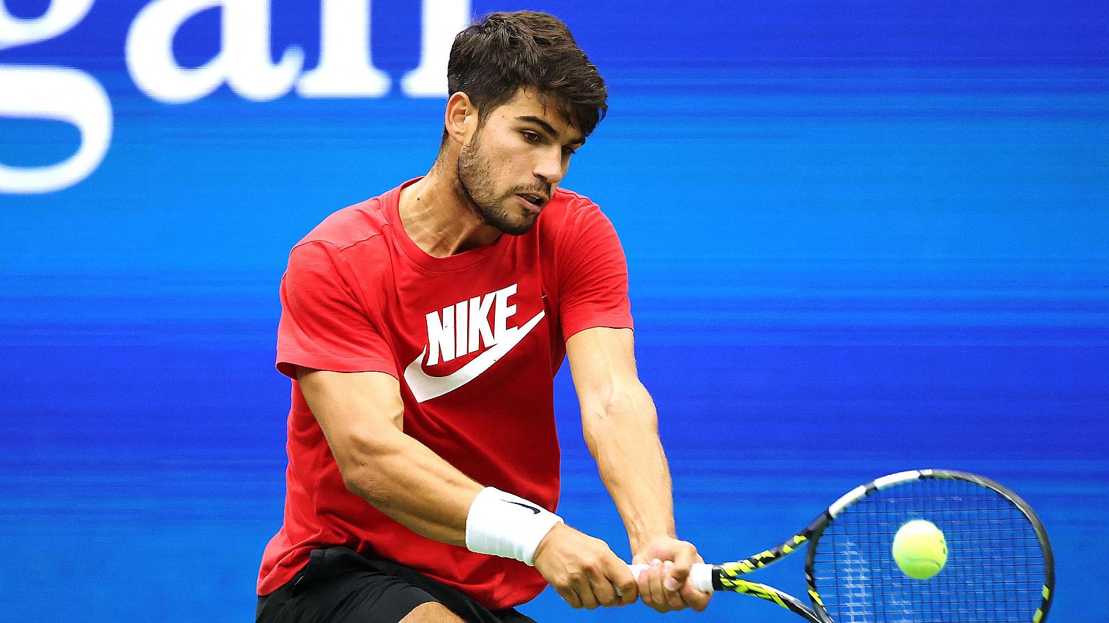 Tenista golpeando una pelota amarilla con su raqueta. Lleva camiseta roja y muñequera blanca. Fondo azul intenso.