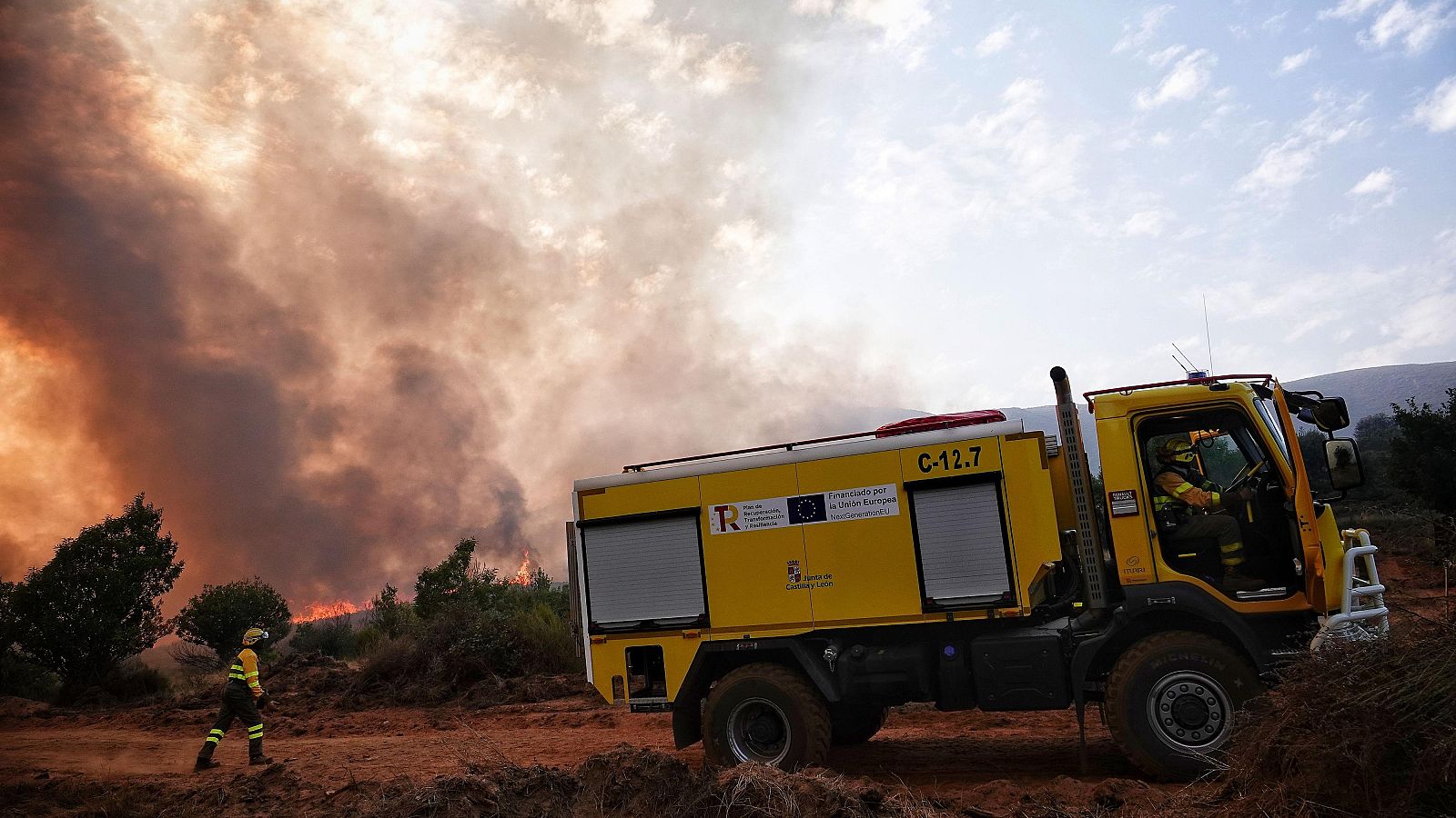 Camión de bomberos (matrícula C-12.7) en un incendio forestal. Dos bomberos, uno conduciendo y otro caminando hacia las llamas.