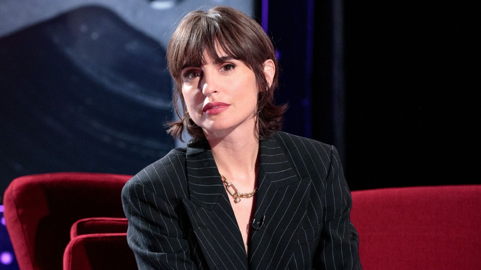 Actriz con pelo corto y castaño oscuro, chaqueta oscura con rayas blancas y collar dorado, sentada en un sofá rojo.