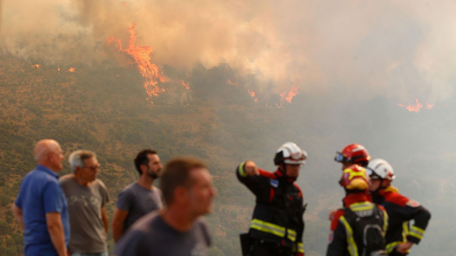 Incendio forestal activo con llamas anaranjadas y densa columna de humo.  Bomberos y civiles observan la situación.