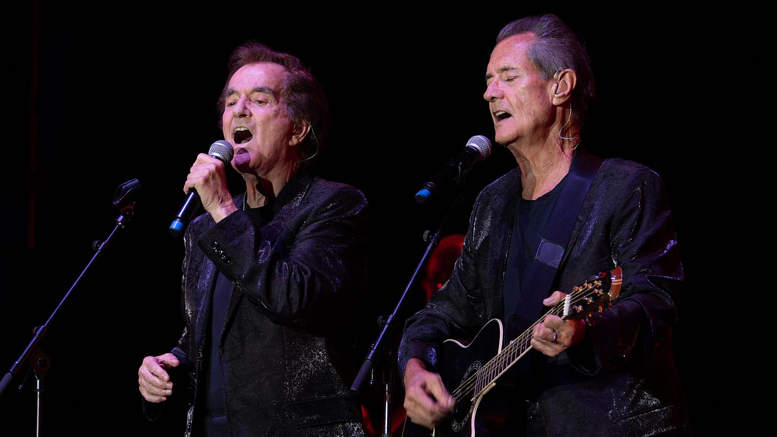 Concierto del Dúo Dinámico: Manuel de la Calva canta con micrófono en mano mientras Ramón Arcusa toca la guitarra electroacústica; ambos con chaquetas oscuras.