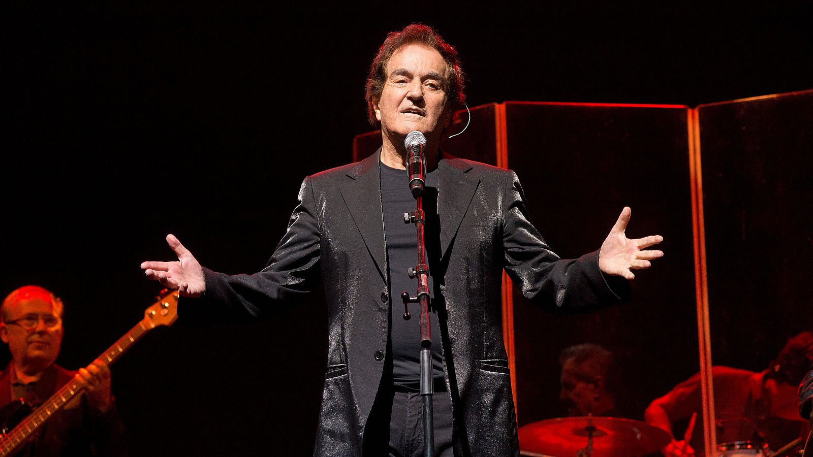 Cantante con chaqueta negra actuando en escenario con iluminación roja, micrófono en mano y brazos extendidos.  Músicos adicionales al fondo.