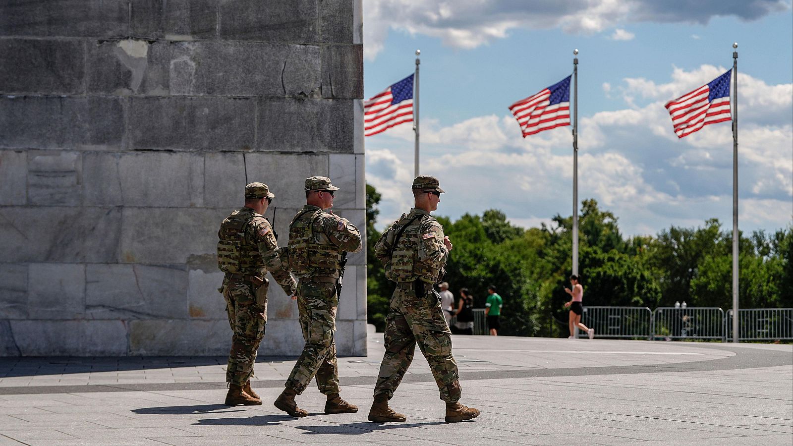 Soldados de la Guardia Nacional patrullan, armados y en uniforme de camuflaje, la base del Monumento a Washington, con banderas estadounidenses ondeando al fondo.