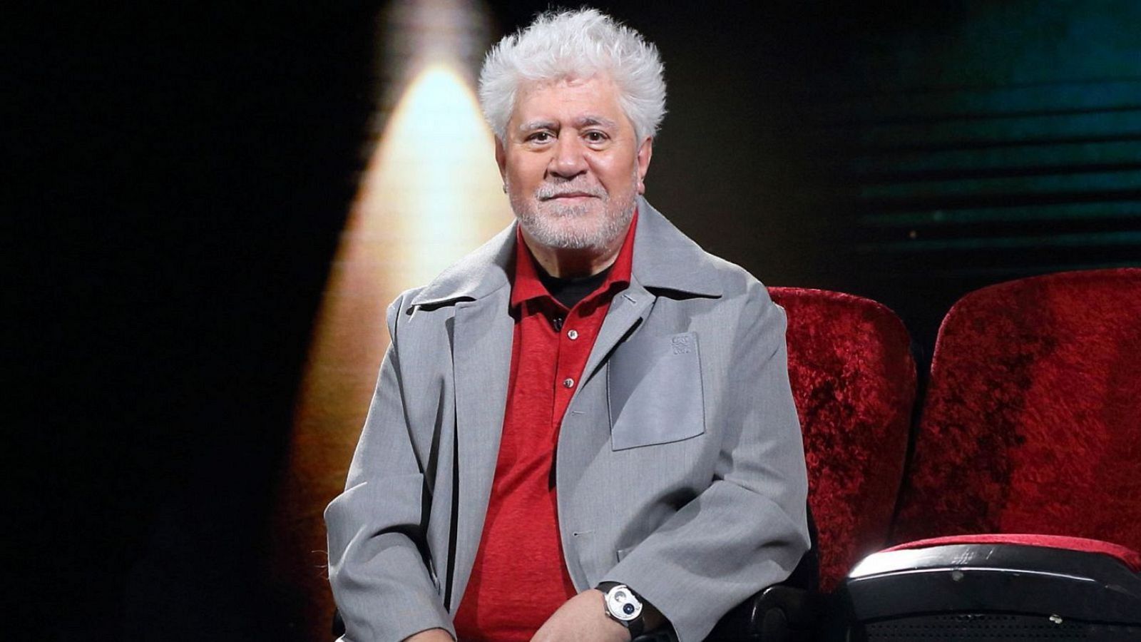 Imagen de archivo de Pedro Almodóvar sentado en una butaca roja, con chaqueta gris y camisa roja. Fondo oscuro.