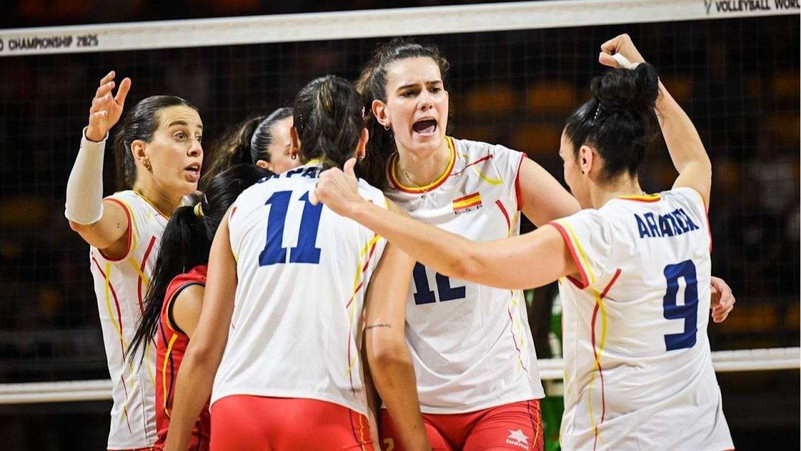 Jugadoras de voleibol, vestidas con uniforme blanco, rojo y amarillo, celebran un punto con abrazos y gestos de alegría. Se ven dorsales 9, 11 y 12.