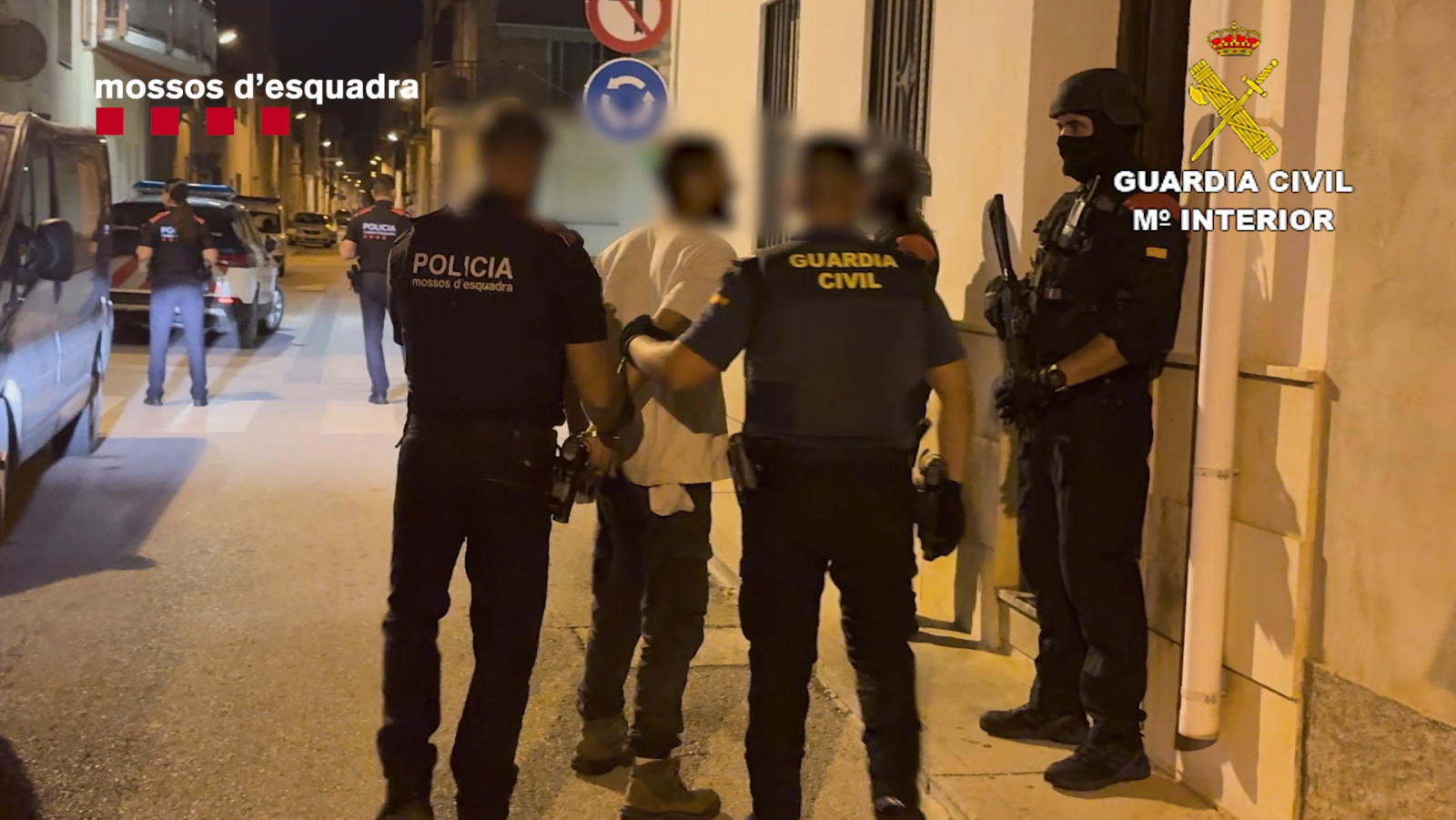 Detención policial en una calle. Dos agentes, Mossos d'Esquadra y Guardia Civil, escoltan a un detenido esposado. Se observan otros agentes y vehículos policiales.