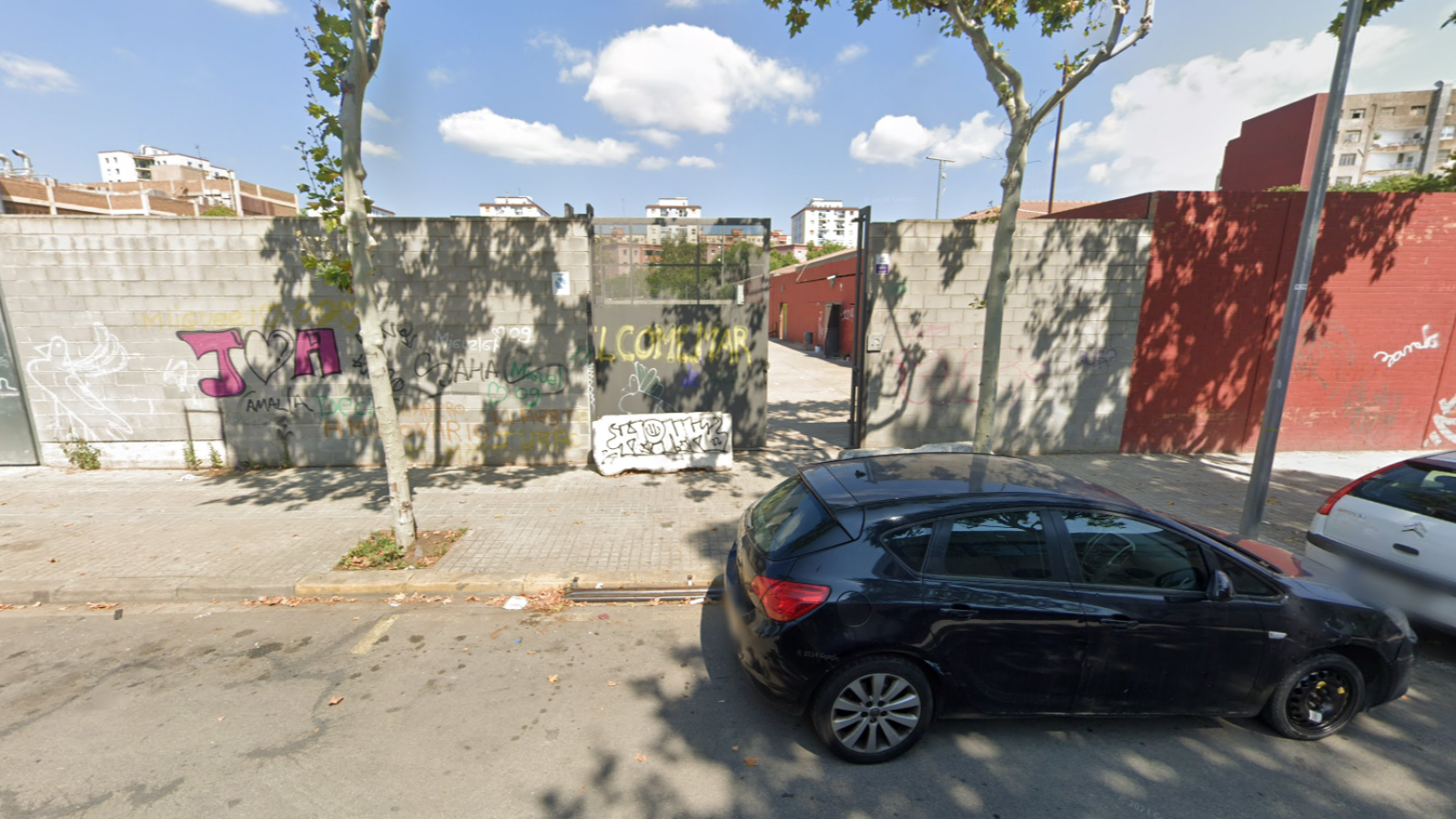 Muro con grafitis ("J" y "A" con corazón, "WELCOME") y puerta metálica.  Coche negro aparcado en la calle.  Vista de Google Maps.