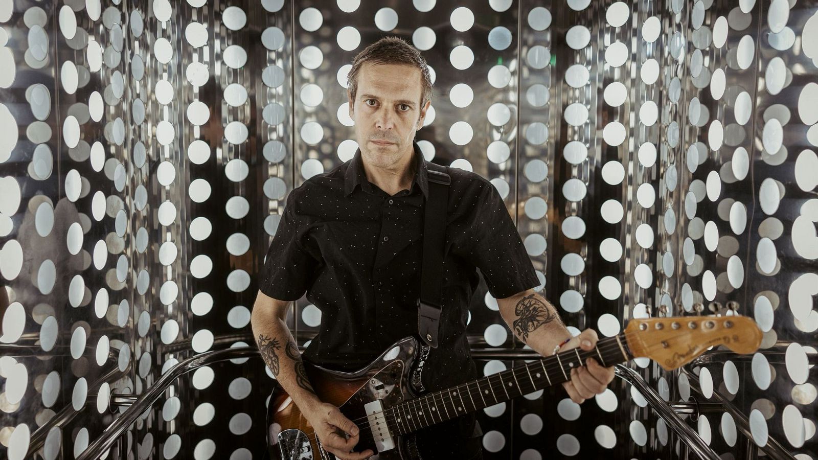 Músico con camisa negra sostiene guitarra en espacio con paredes circulares.  Expresión seria, tatuajes visibles en brazos.