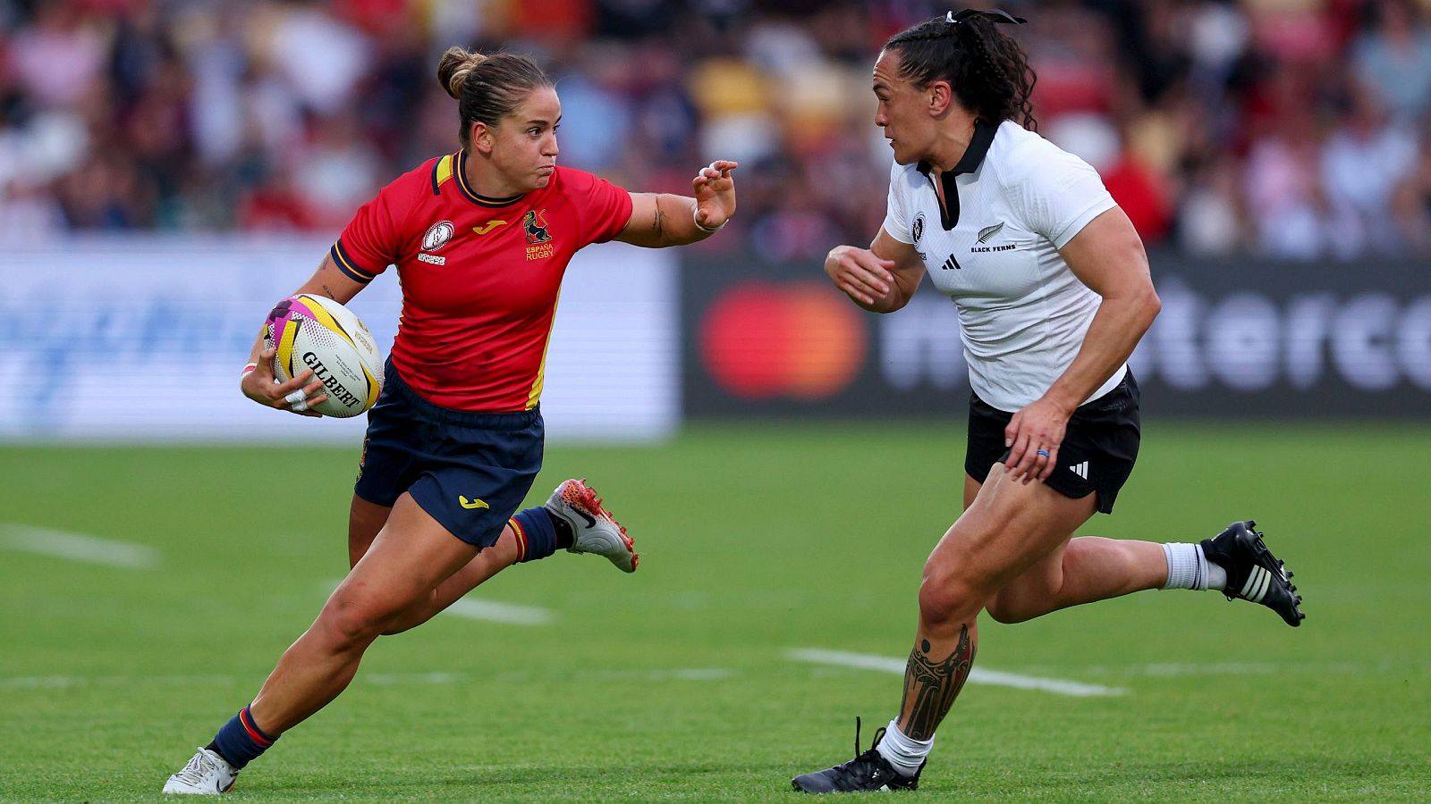 Jugadora de rugby española con equipación roja corre con el balón, perseguida por una rival neozelandesa (camiseta negra y blanca).  Intensidad en el juego.