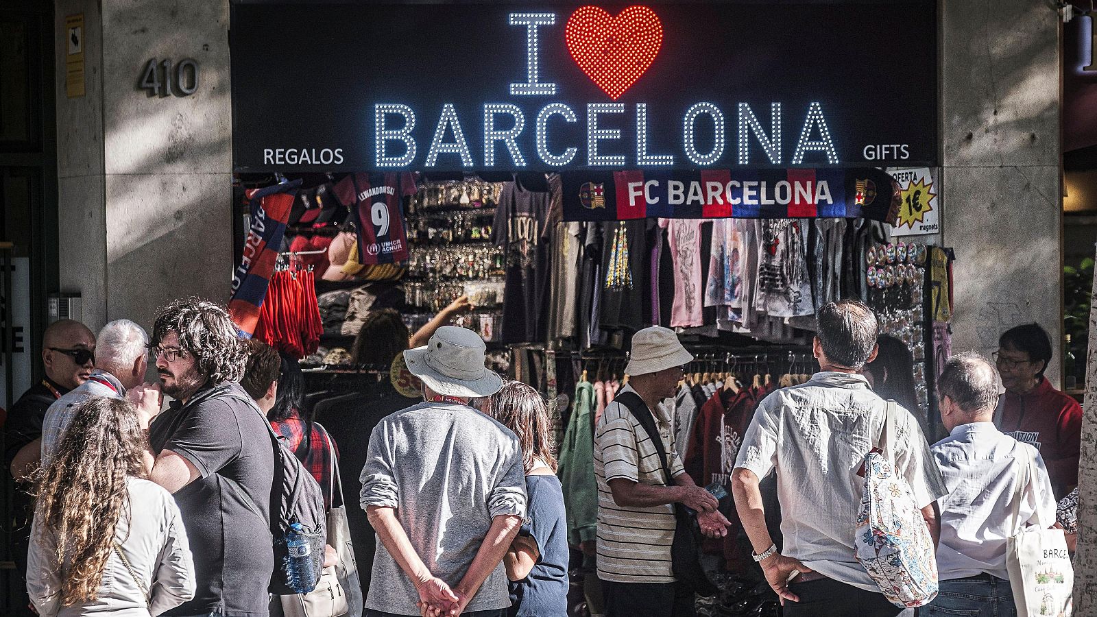 Turistas observan souvenirs en una tienda de Barcelona con un cartel luminoso "I ♥ BARCELONA", incluyendo artículos con motivos deportivos.