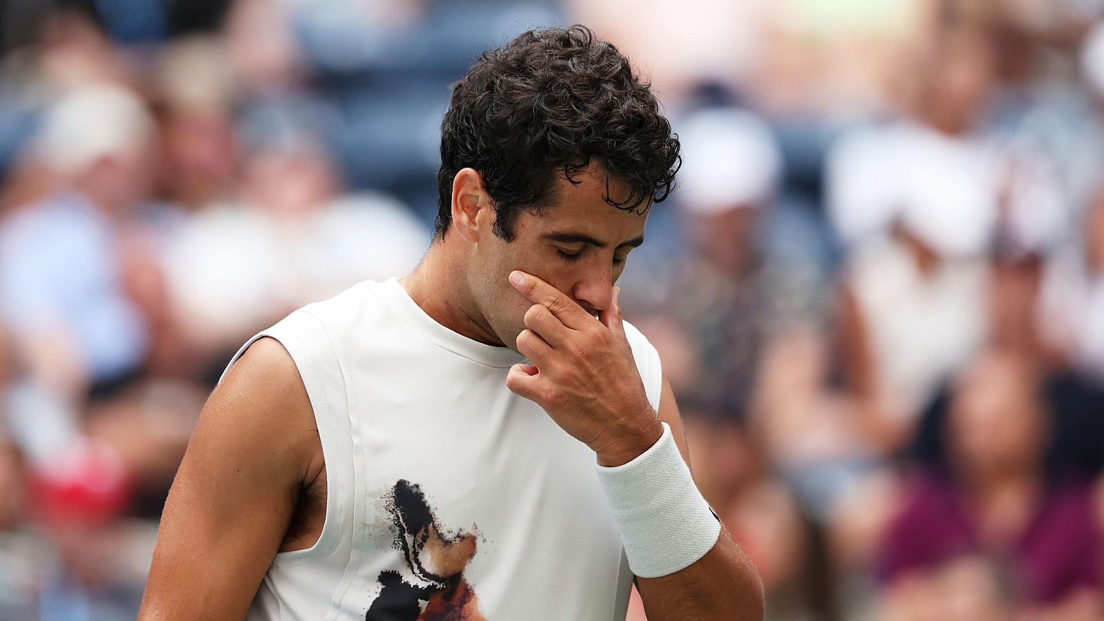 Tenista con camiseta blanca sin mangas y muñequera, gesto de concentración o cansancio con la mano en la cara. Fondo borroso con público.