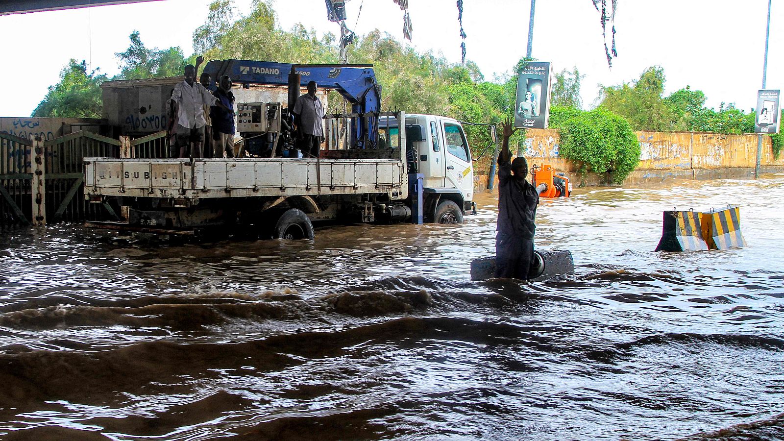 Inundación: Personas en un camión con grúa, parcialmente sumergido, y otra persona en el agua pidiendo auxilio.  Agua turbia cubre gran parte de la escena.