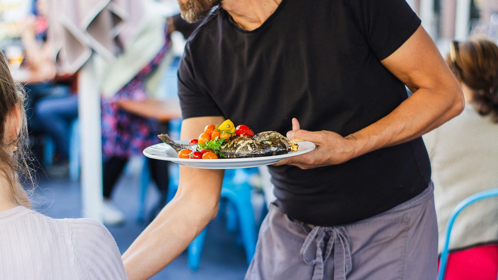 Camarero sirve pescado a la plancha con guarnición a clienta en terraza concurrida.  Ambiente de restaurante con mesas y sillas azules.