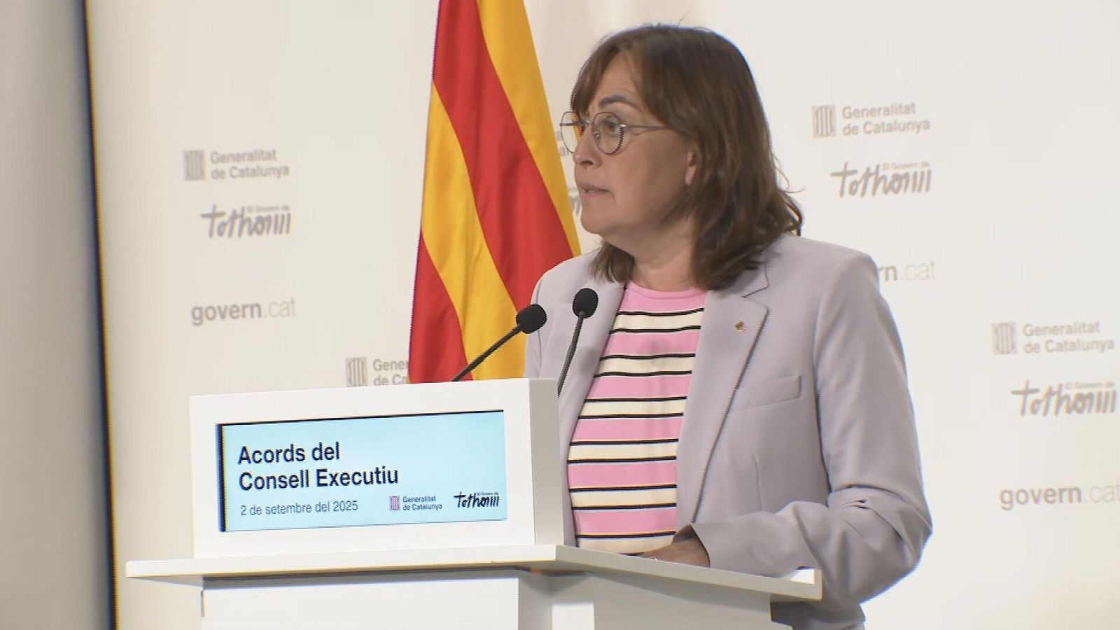Rueda de prensa: mujer con gafas y chaqueta clara detrás de un atril con la información "Acords del Consell Executiu", fecha 2 de septiembre de 2025 y la bandera de Cataluña.