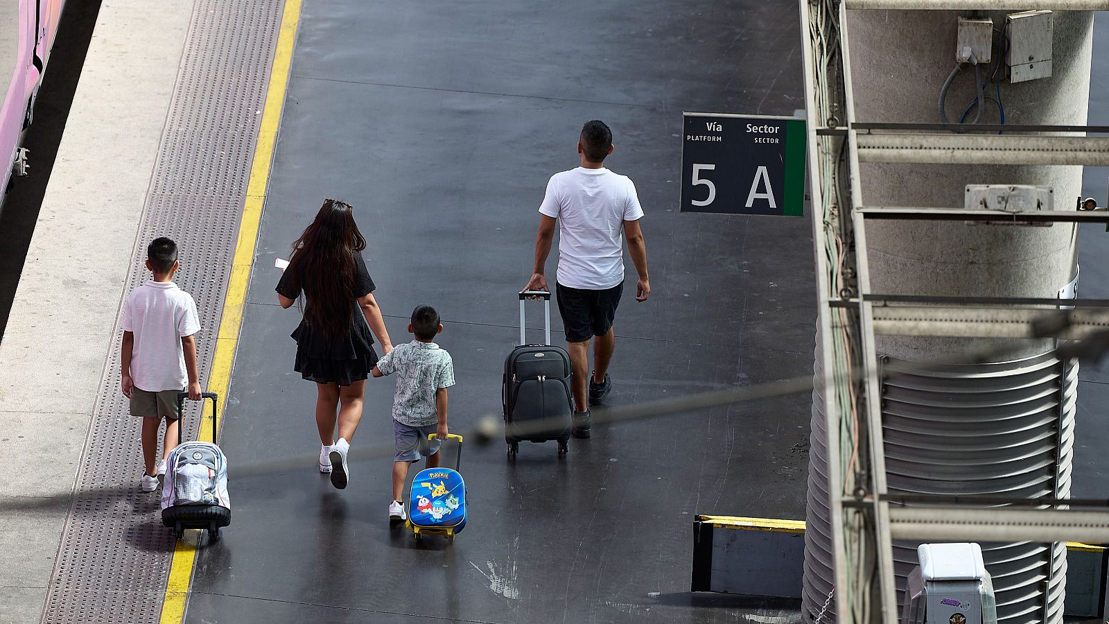Familia con niños y equipaje camina por andén de estación, esperando tren.  Se observa un tren rosa y señalización de andén.