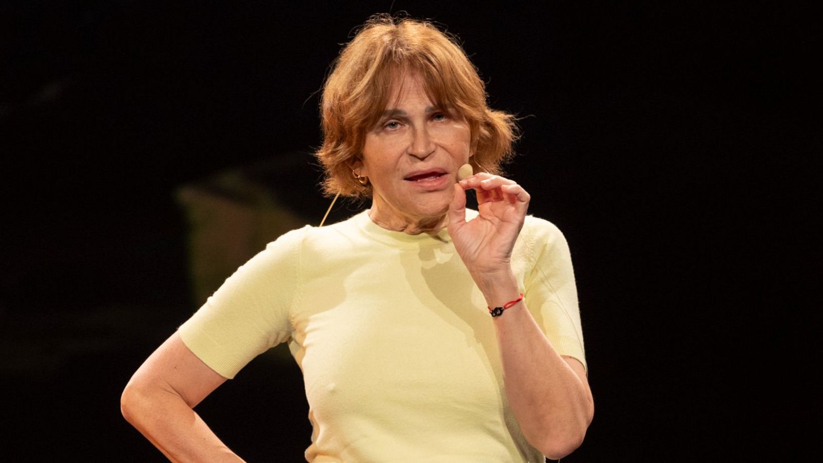 Actriz con pelo corto rojizo y camiseta amarilla, gesticulando con la mano derecha cerca de la boca. Fondo oscuro.