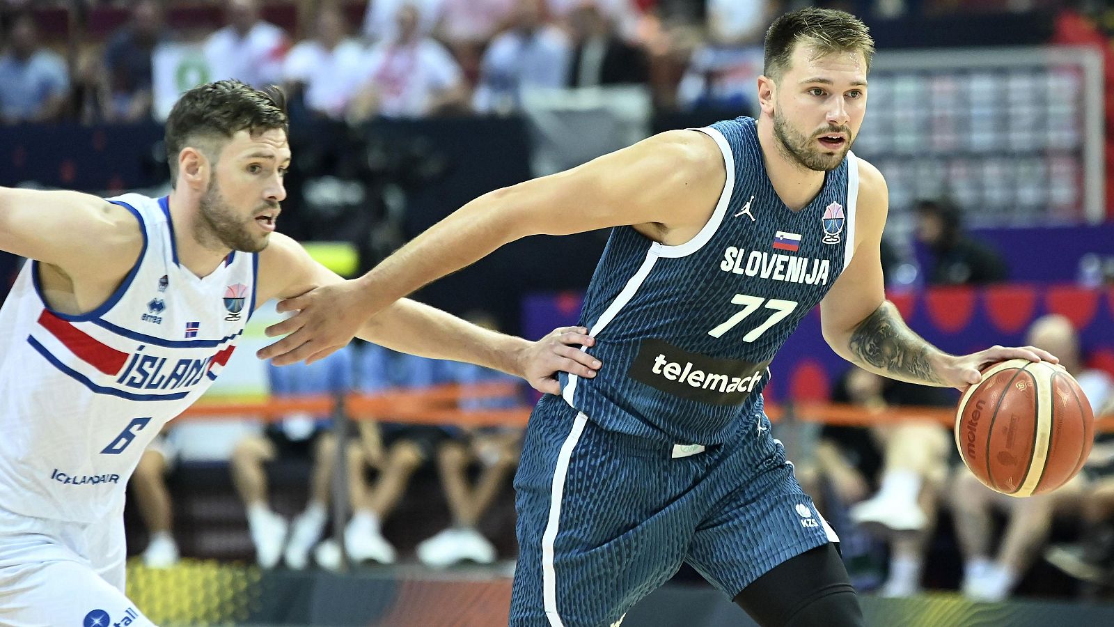 Partido de baloncesto: jugador islandés (#6, camiseta blanca con franjas) defiende a jugador esloveno (#77, camiseta azul) con el balón.  Acción deportiva.