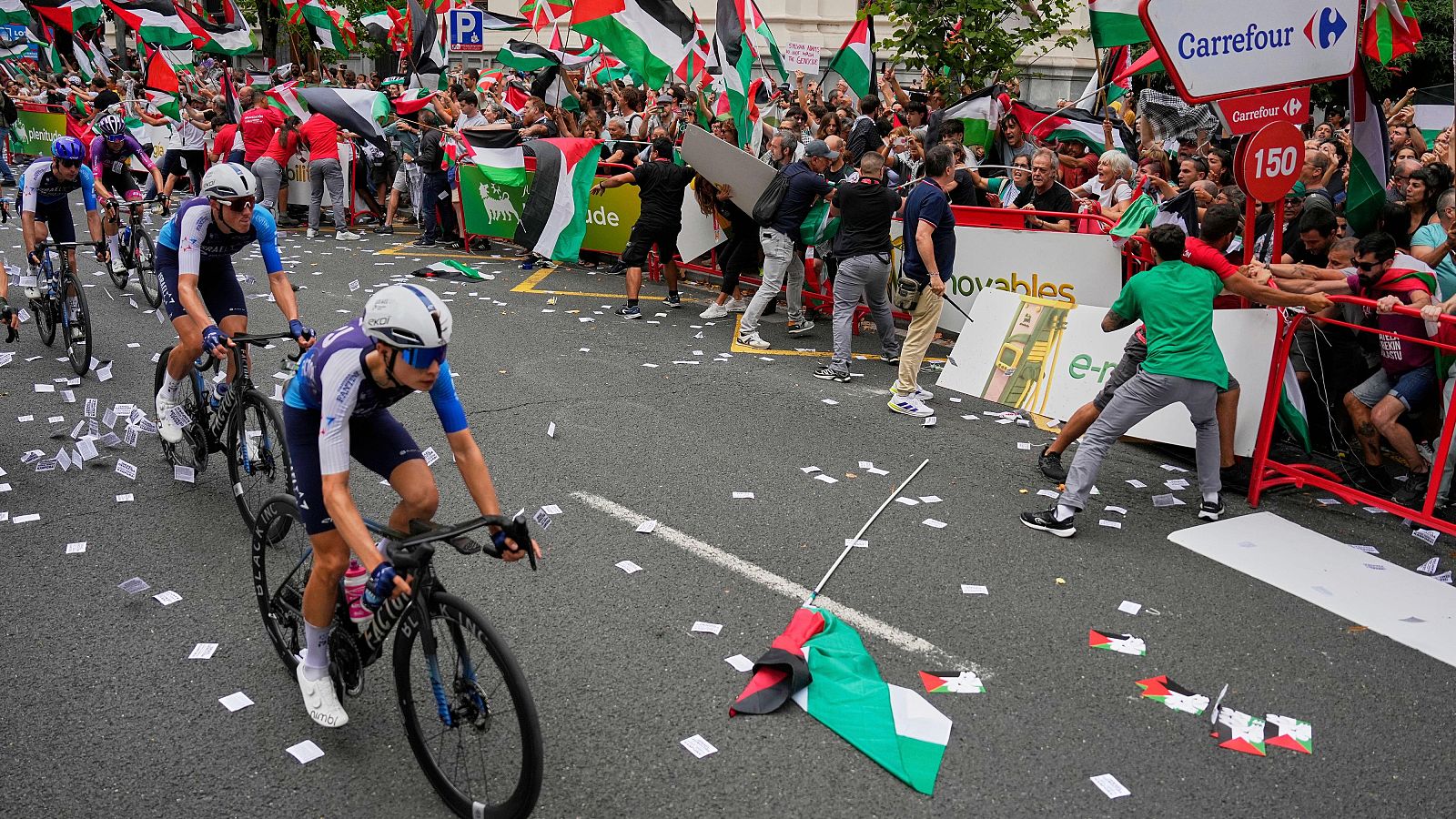 Ciclistas en carrera, multitud con banderas palestinas y personas intentando romper vallas de seguridad.  Ambiente tenso.