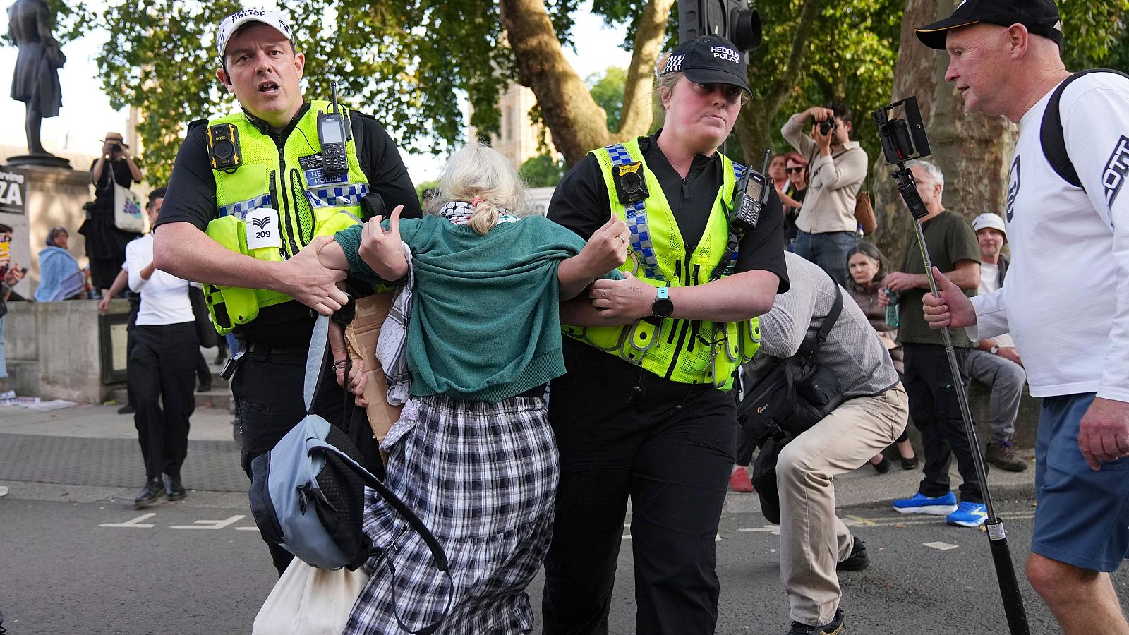 Policías detienen a una manifestante de cabello rubio/grisáceo con camisa verde y falda a cuadros.  Otros observan y graban la escena con teléfonos y un palo selfie.