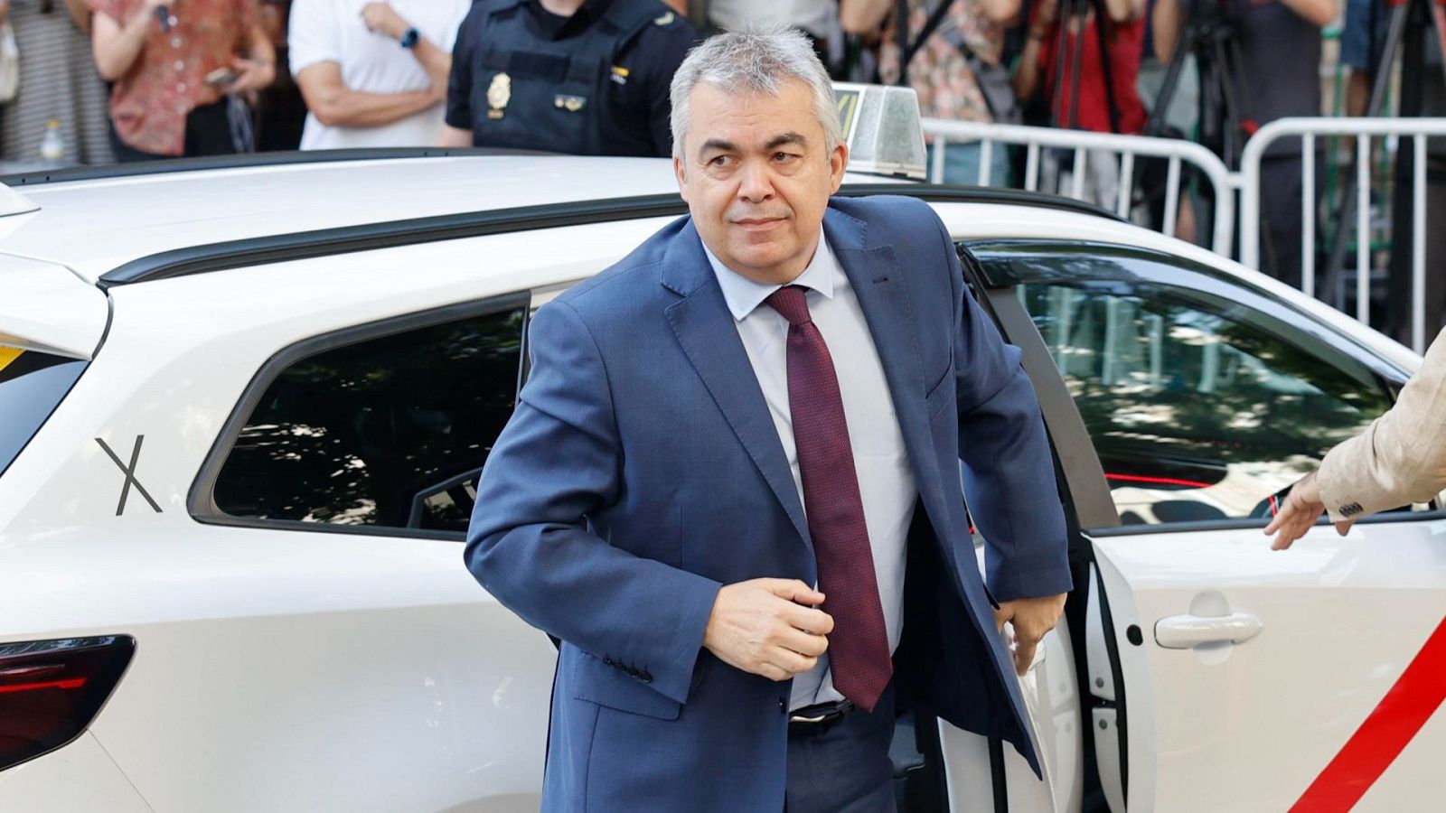 Un hombre en traje azul marino y corbata granate sale de un coche blanco con franja roja.  Parece llegar a un edificio, observado por un grupo de personas.