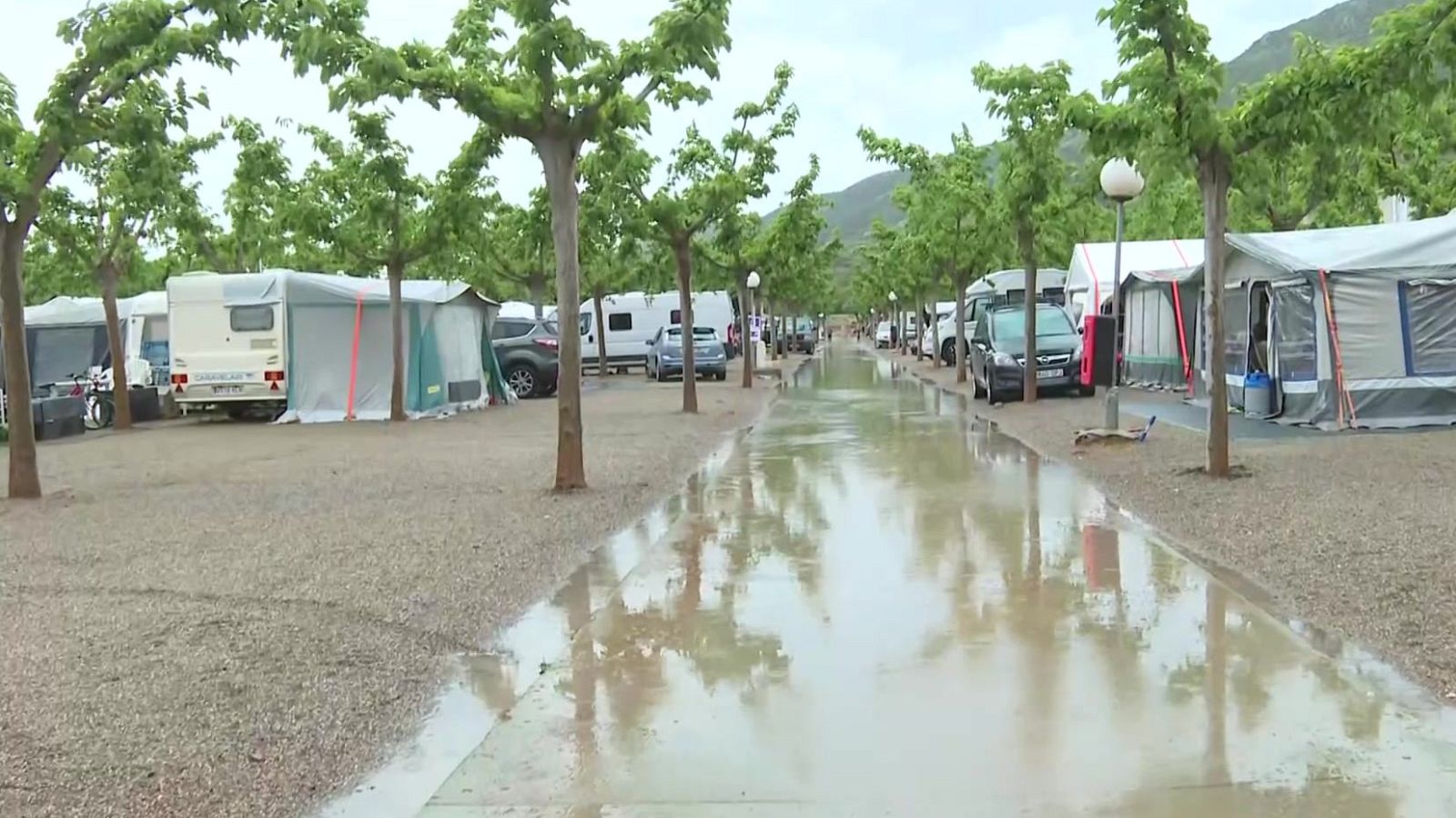 Inundación en un camping: camino central convertido en charco, reflejando el cielo y árboles.  Caravanas, tiendas y coches aparcados en zona de gravilla.