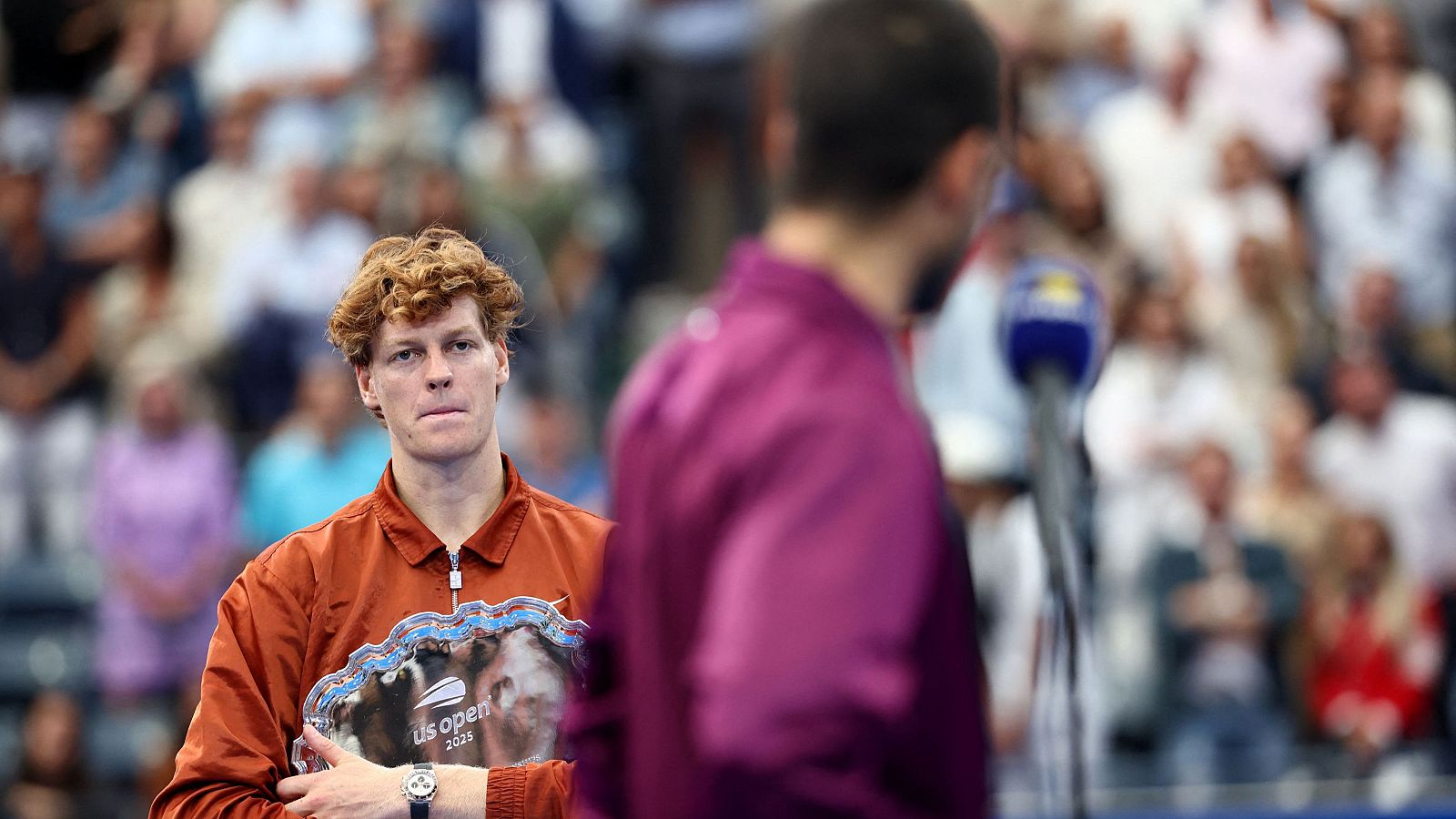Tenista con chaqueta marrón rojiza y premio, expresión seria, tras una competición.  Otro tenista, con chaqueta morada, de fondo, da declaraciones a la prensa.