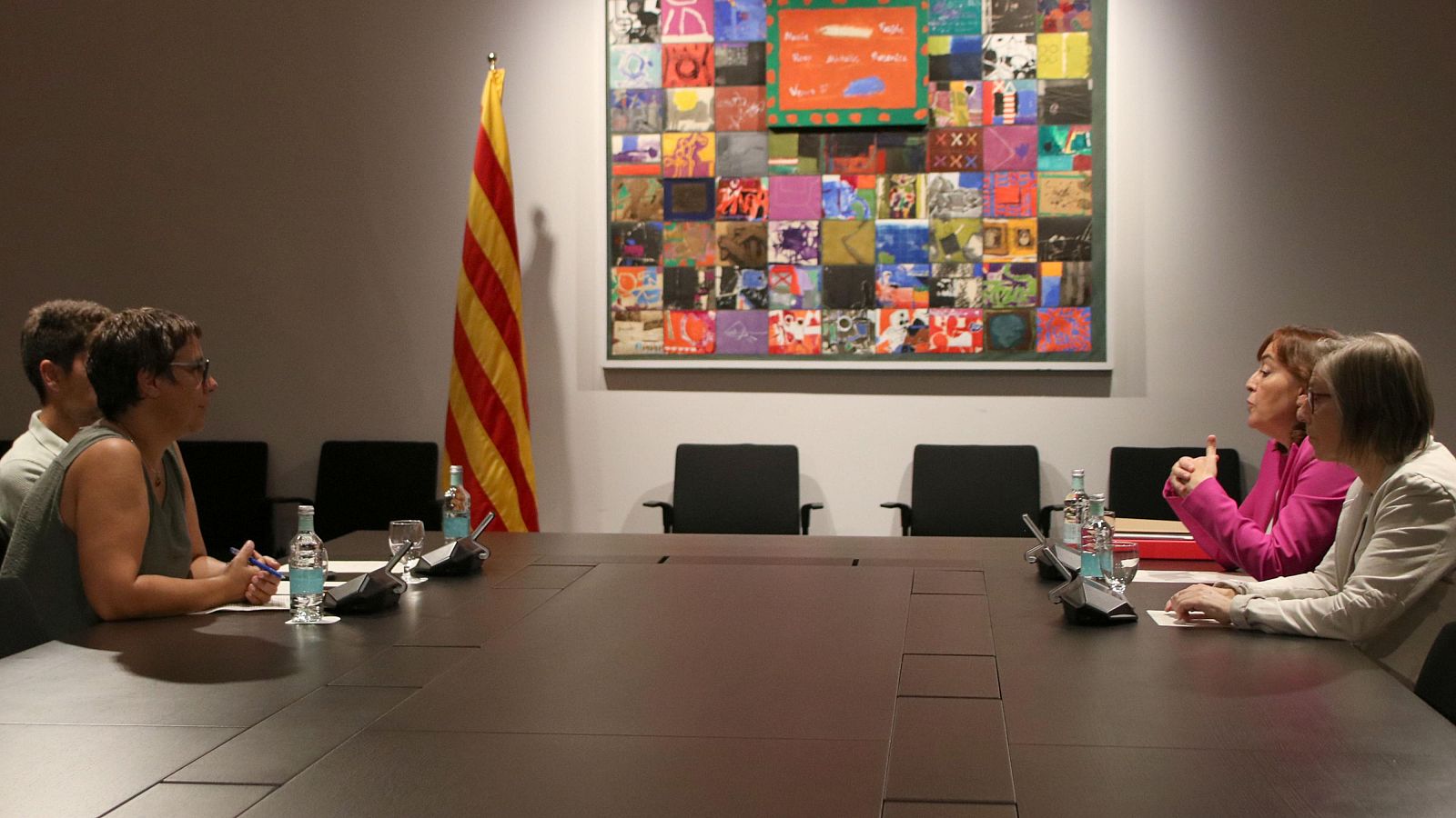 Reunión formal en el Palau de la Generalitat. Cuatro personas alrededor de una mesa con micrófonos y agua.  Se observa una bandera catalana y un cuadro en la pared.
