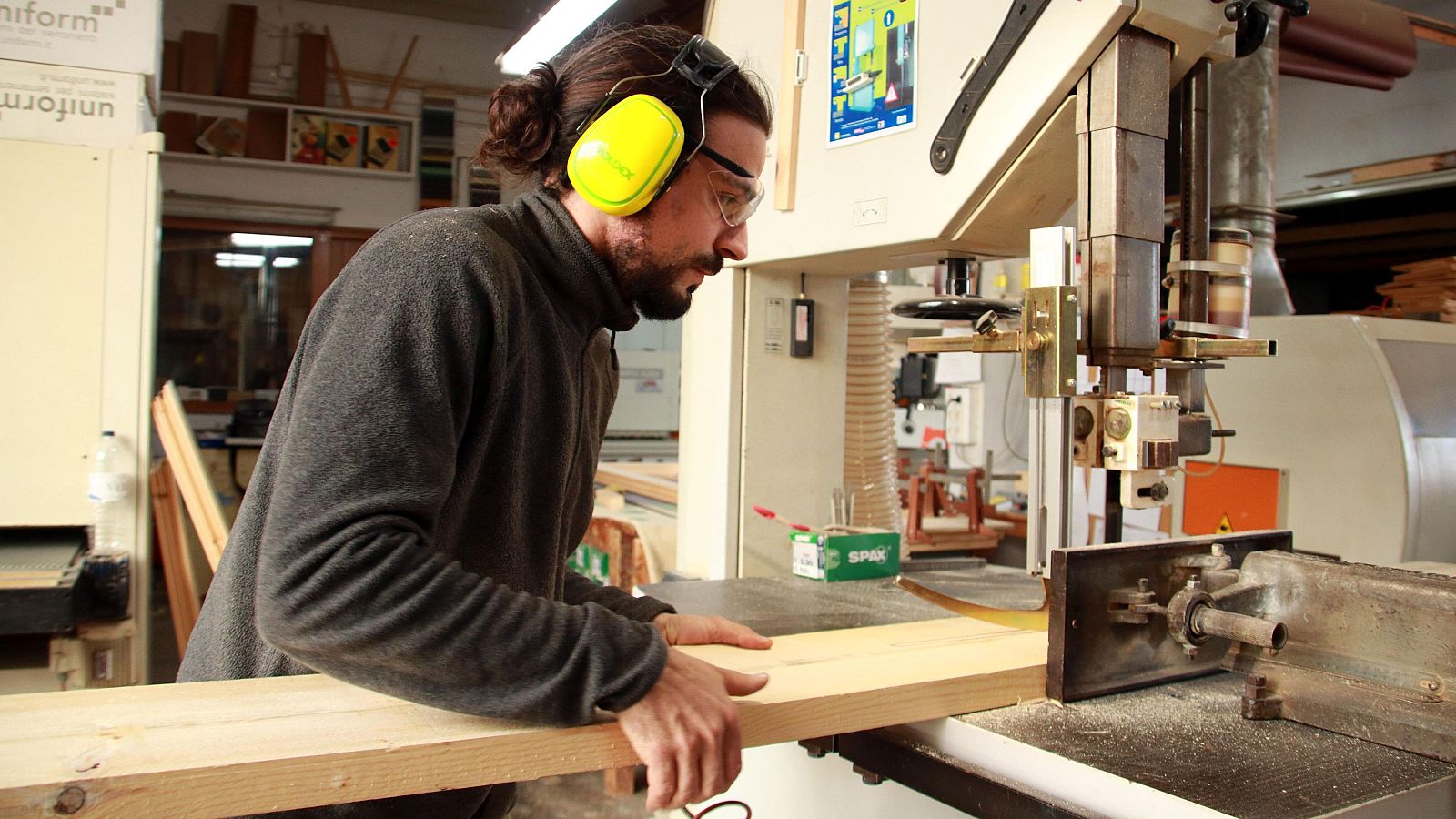Carpintero trabajando con sierra de cinta, usando protección auditiva y visual.  Imagen relacionada con la demanda de profesionales en oficios.