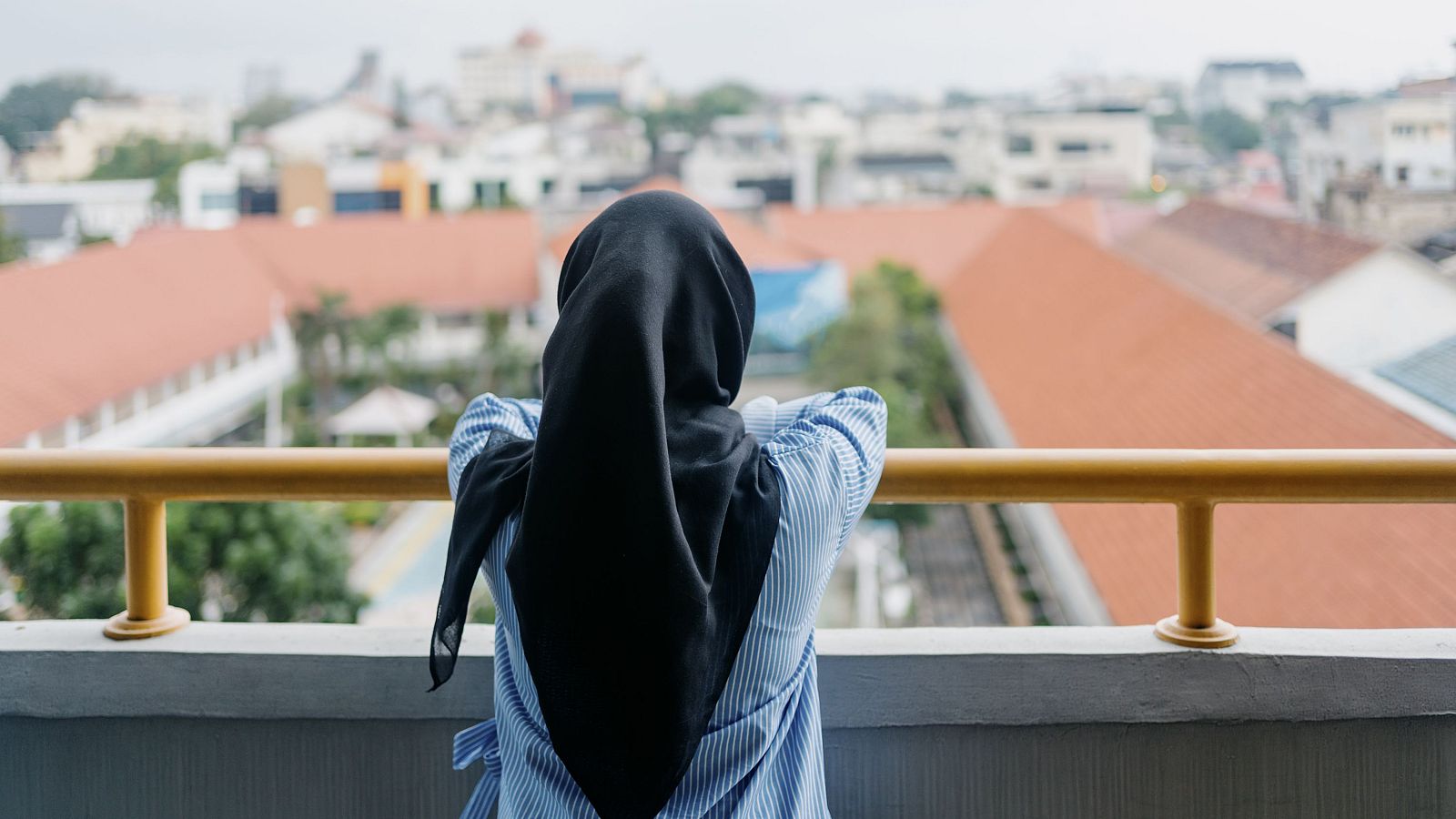 Joven con hiyab negro y camisa azul claro a rayas, apoyada en una barandilla dorada, observa un paisaje urbano difuso.