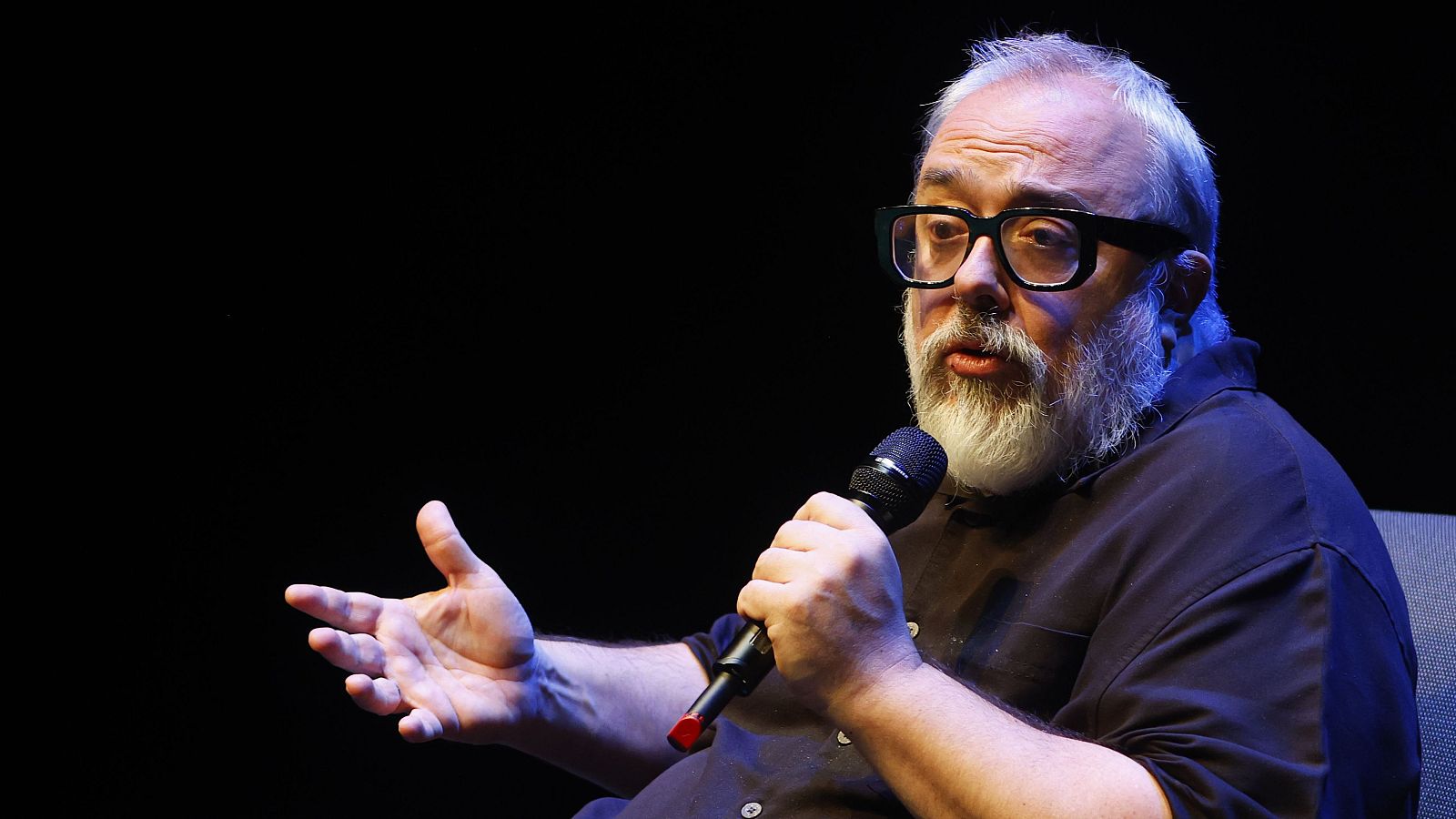 El director Álex de la Iglesia, con gafas y barba, habla con un micrófono en mano en un evento con iluminación tenue.