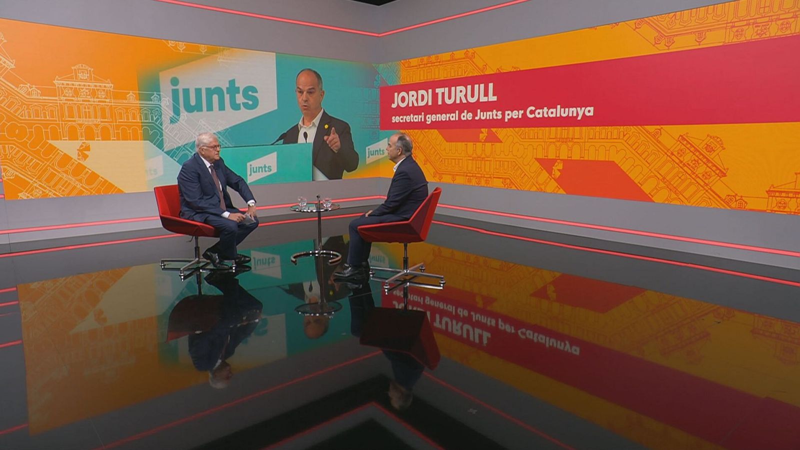 Entrevista a Jordi Turull a 'Aquí Parlem'.  Assegut en cadira vermella, amb vestimenta formal.  Pantalla de fons amb el seu nom i logotip del partit.