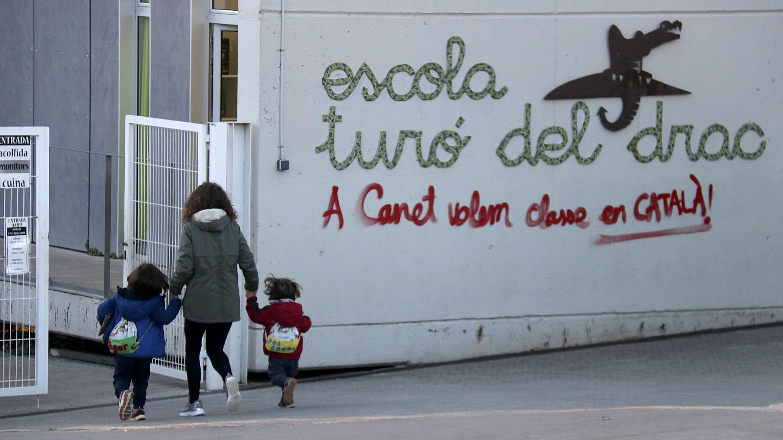 Mujer con dos niños pequeños entrando en una escuela. Grafiti en la fachada reclama clases en catalán.  Escena cotidiana con reivindicación lingüística.