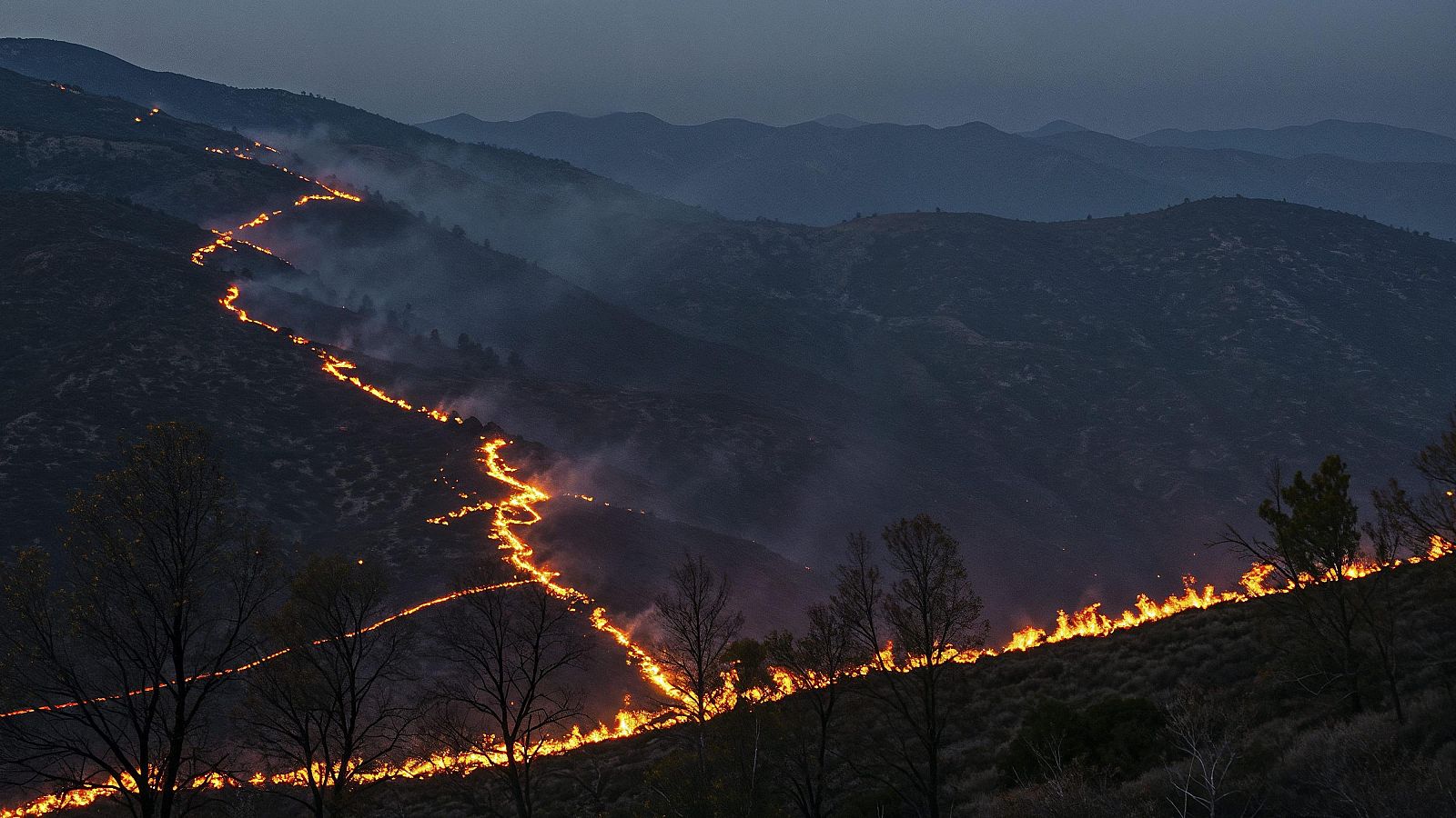 Incendio forestal nocturno en ladera montañosa. Llamas y humo se aprecian con claridad sobre un fondo oscuro.