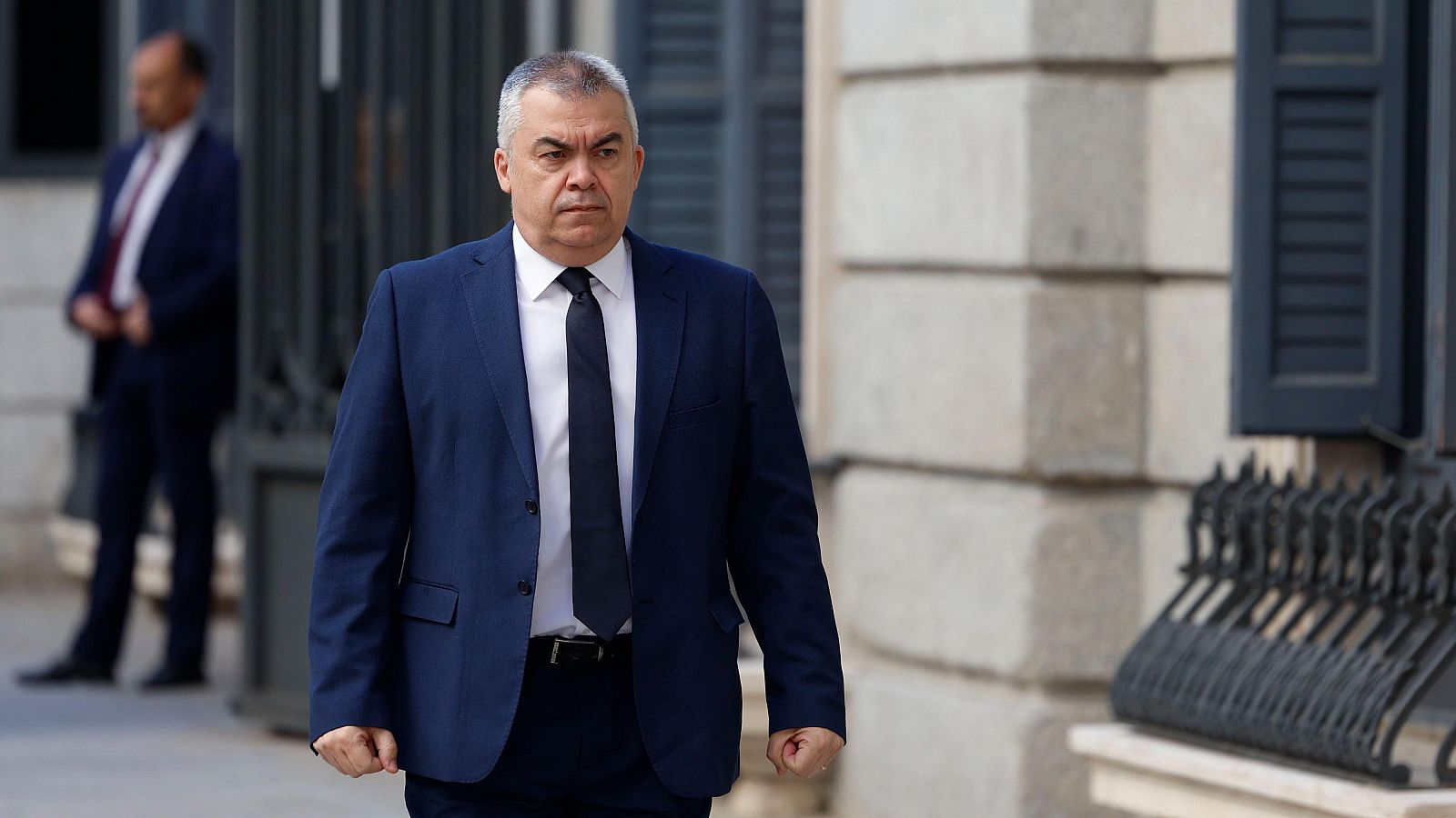 Hombre en traje azul marino camina por la calle con expresión seria; al fondo, otro hombre con traje y un edificio.  Imagen ambientada en entorno urbano.