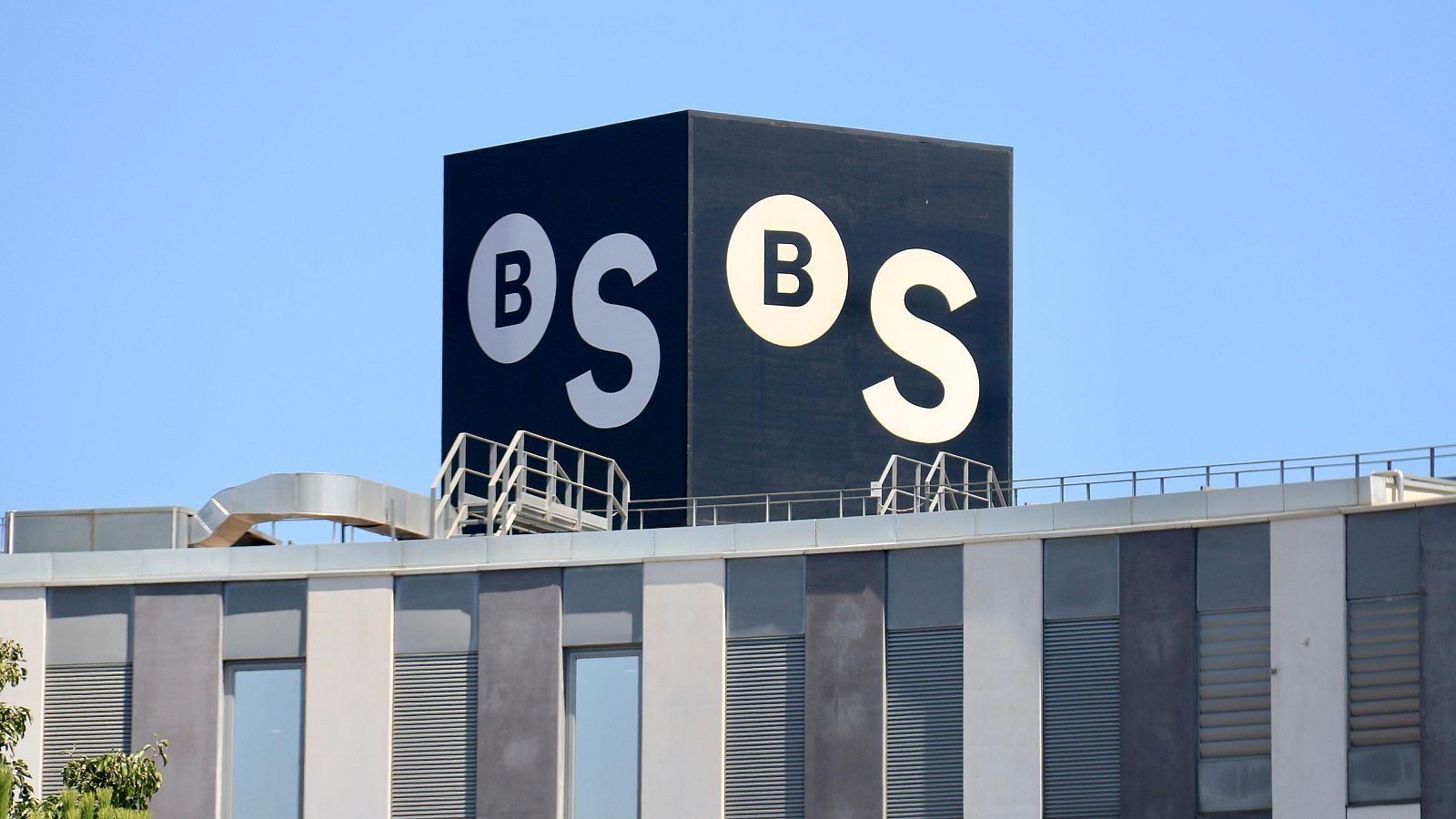 Cubo negro con logotipo en azotea de edificio moderno de fachada gris y blanca.  Escalera metálica visible. Cielo despejado.