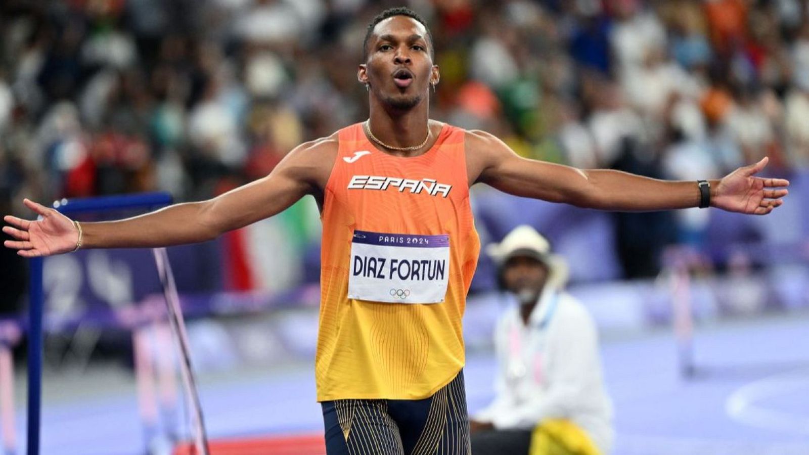 Atleta español con camiseta naranja y amarilla, logo París 2024 y la palabra "ESPAÑA", celebra con los brazos extendidos en un estadio ante un público borroso.