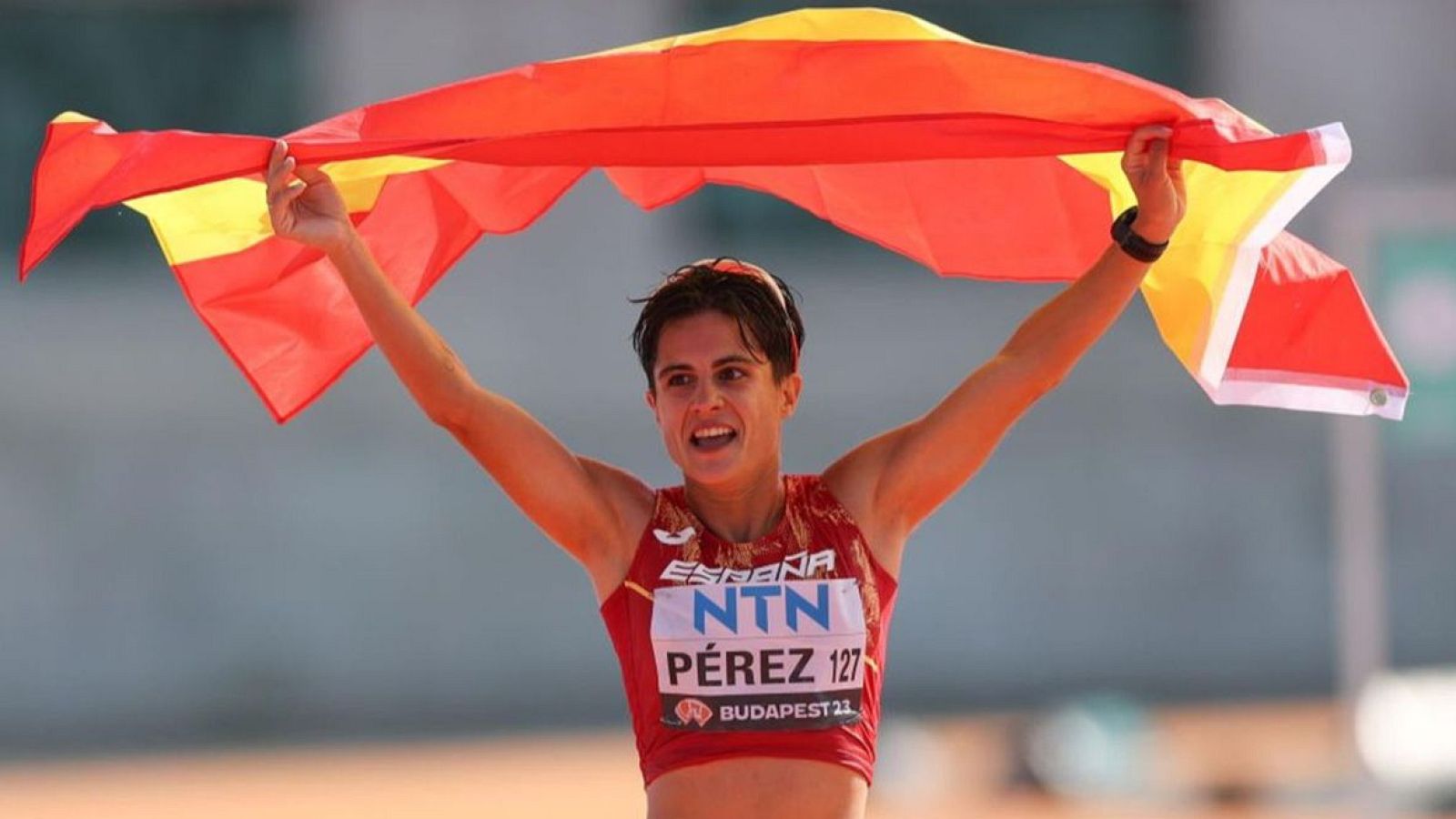 Atleta con camiseta roja y blanca, dorsal 127, celebra victoria ondeando bandera española.  Inscripción "BUDAPEST 23" en la camiseta.