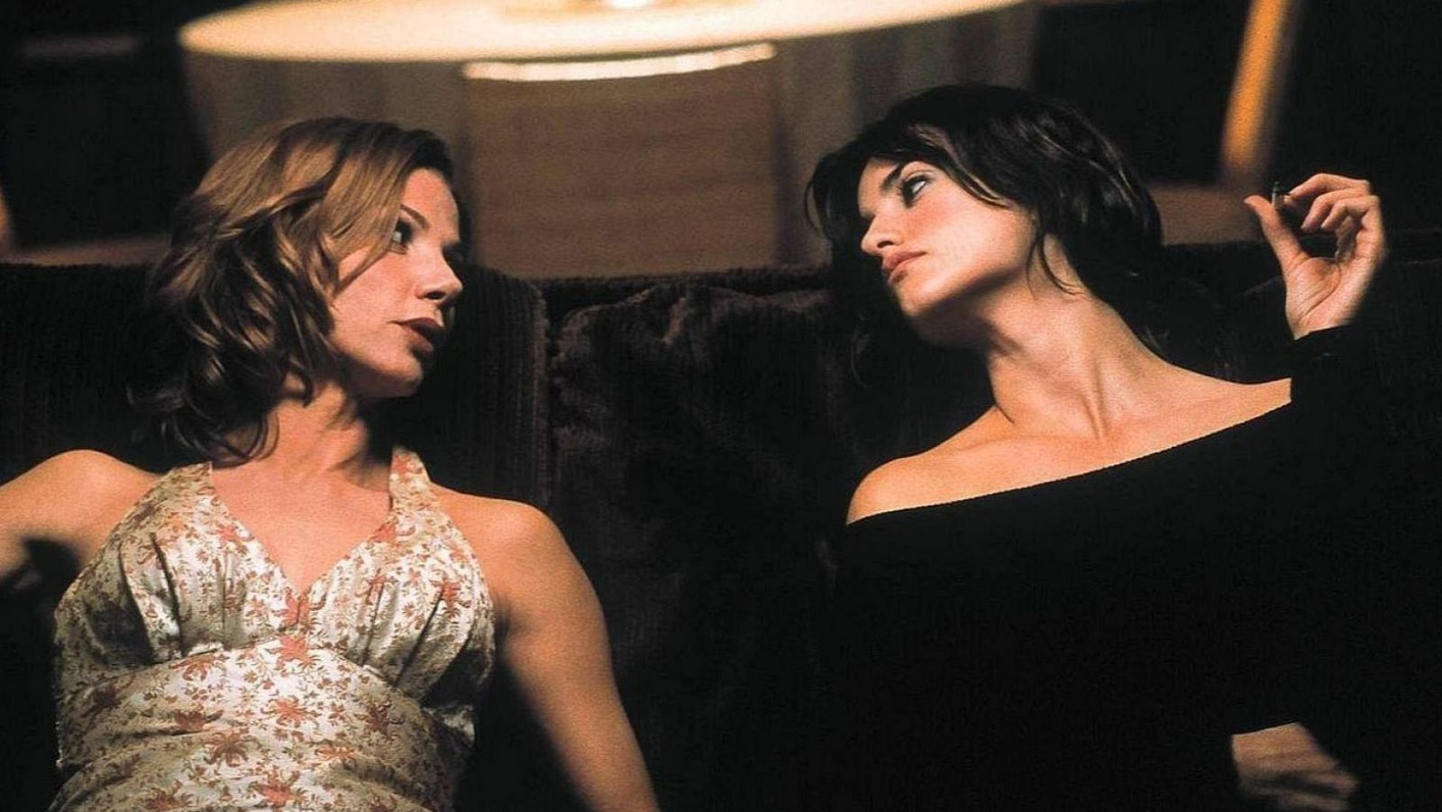 Dos mujeres, una con top floral y otra con top negro, conversan en un espacio oscuro.  Una de ellas parece sostener un cigarrillo.