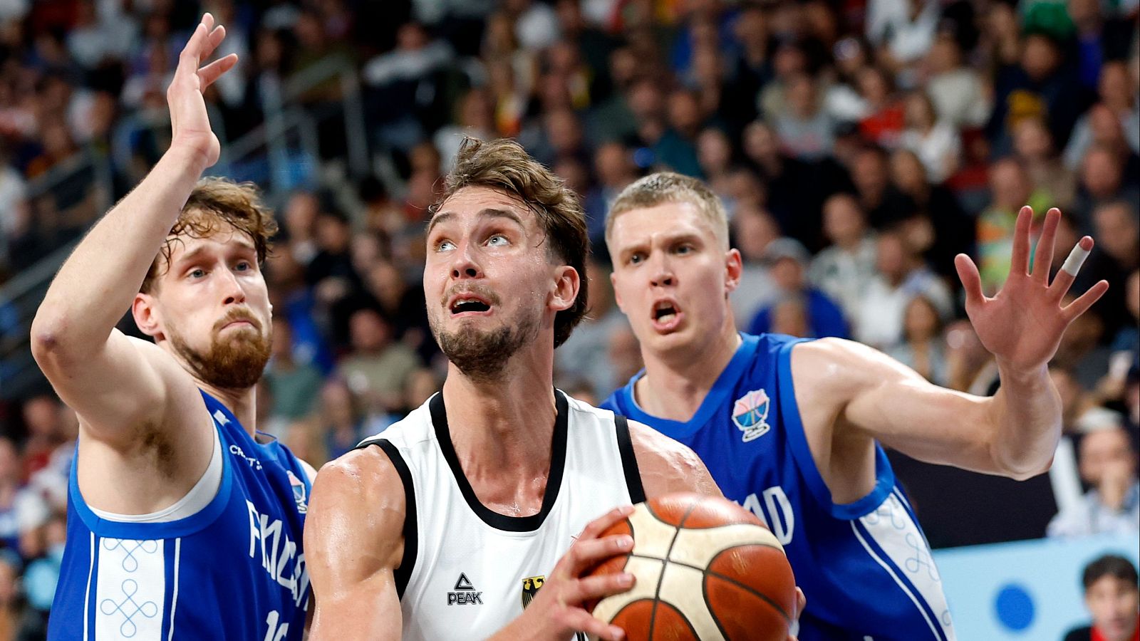 Jugador de baloncesto con balón en mano, rodeado de defensores, concentrado en la jugada.  Público en las gradas.