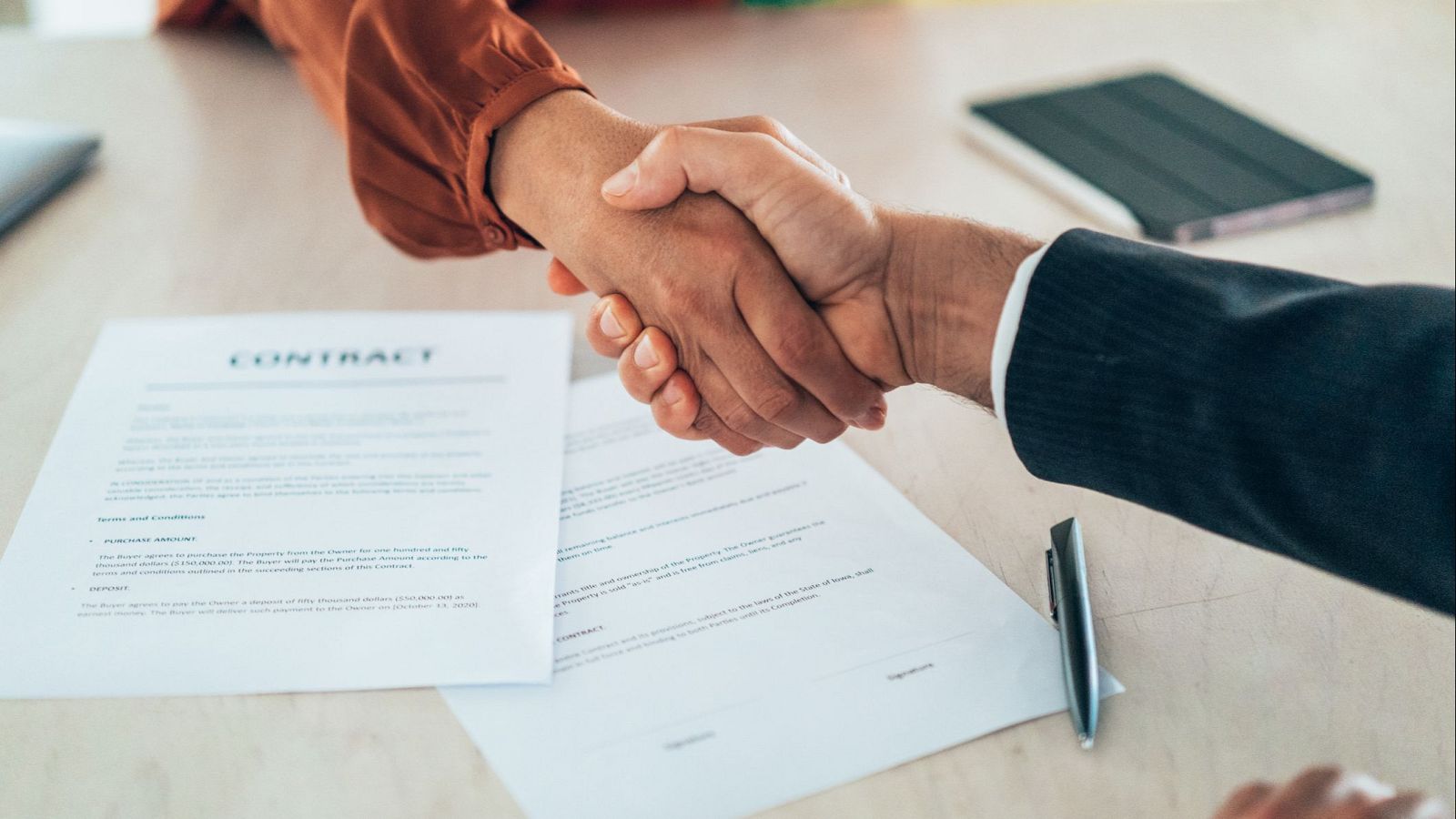 Acuerdo formalizado: dos personas se dan la mano sobre documentos, incluyendo un contrato, un bolígrafo y una tablet.