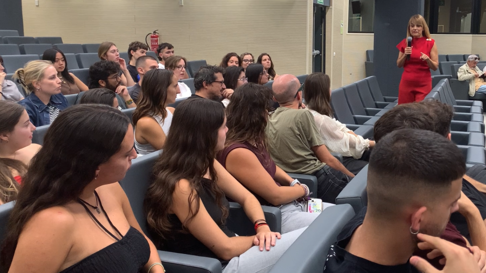 Periodista imparte charla a estudiantes, usando micrófono y gesticulando.  Público atento en sala de conferencias.