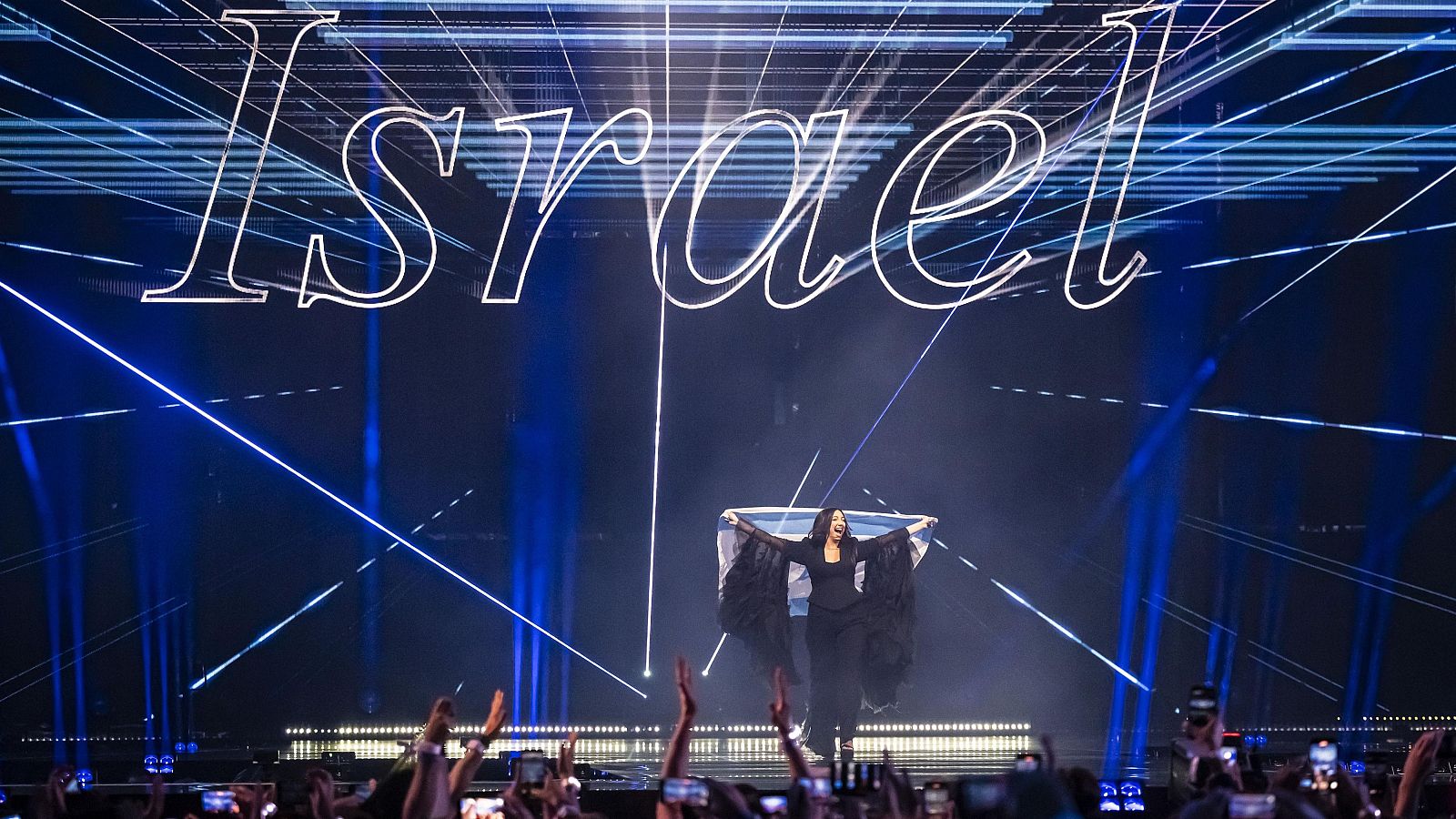 La representant d'Israel a Eurovisió, amb la bandera, sobre l'escenari amb projecció de "Israel" al fons i el públic gravant.