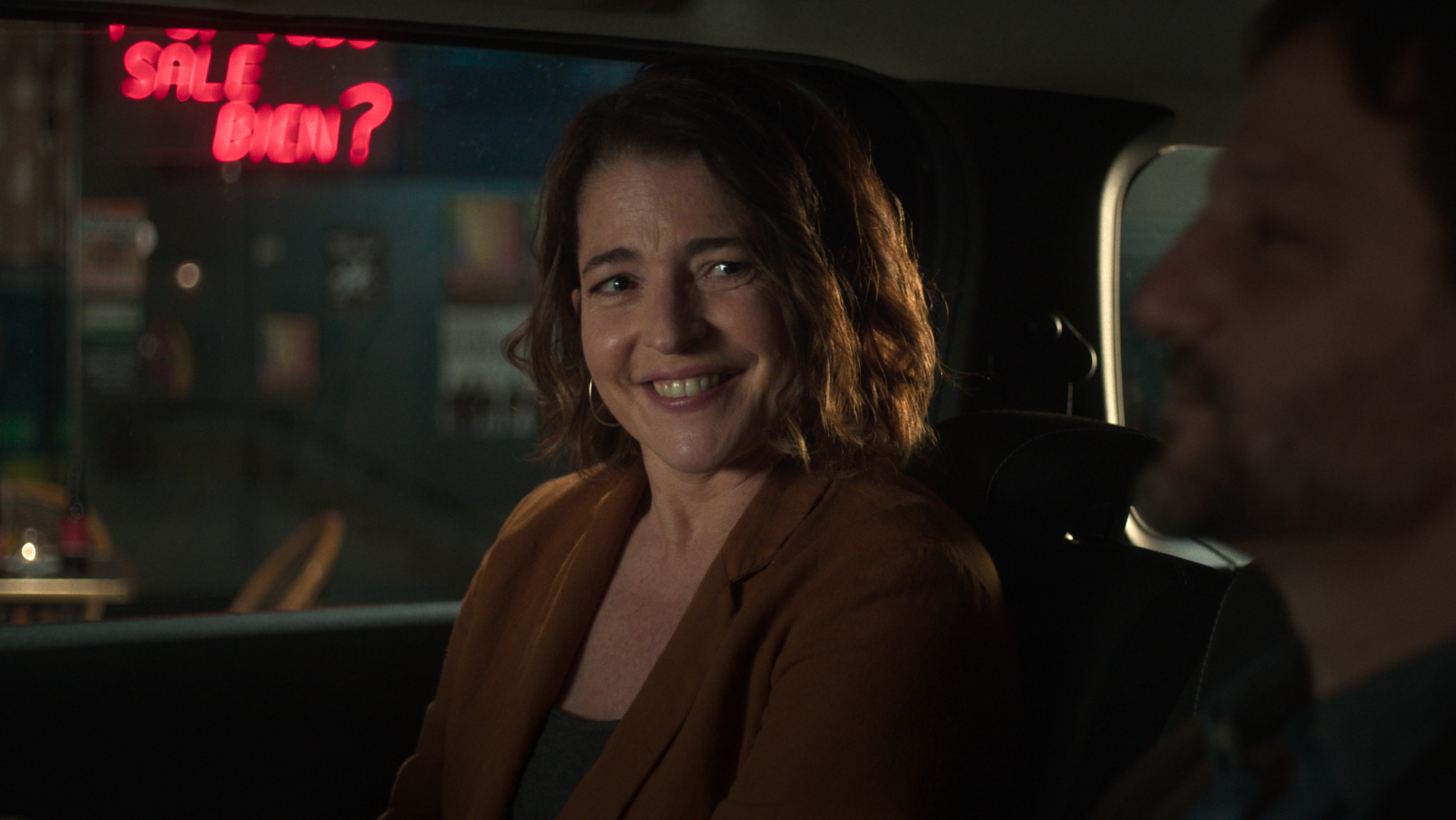 Actriz sonriendo en coche, chaqueta marrón y pendientes.  Se ve un letrero de neón rojo y parte de otra persona. Escena con ambiente positivo.