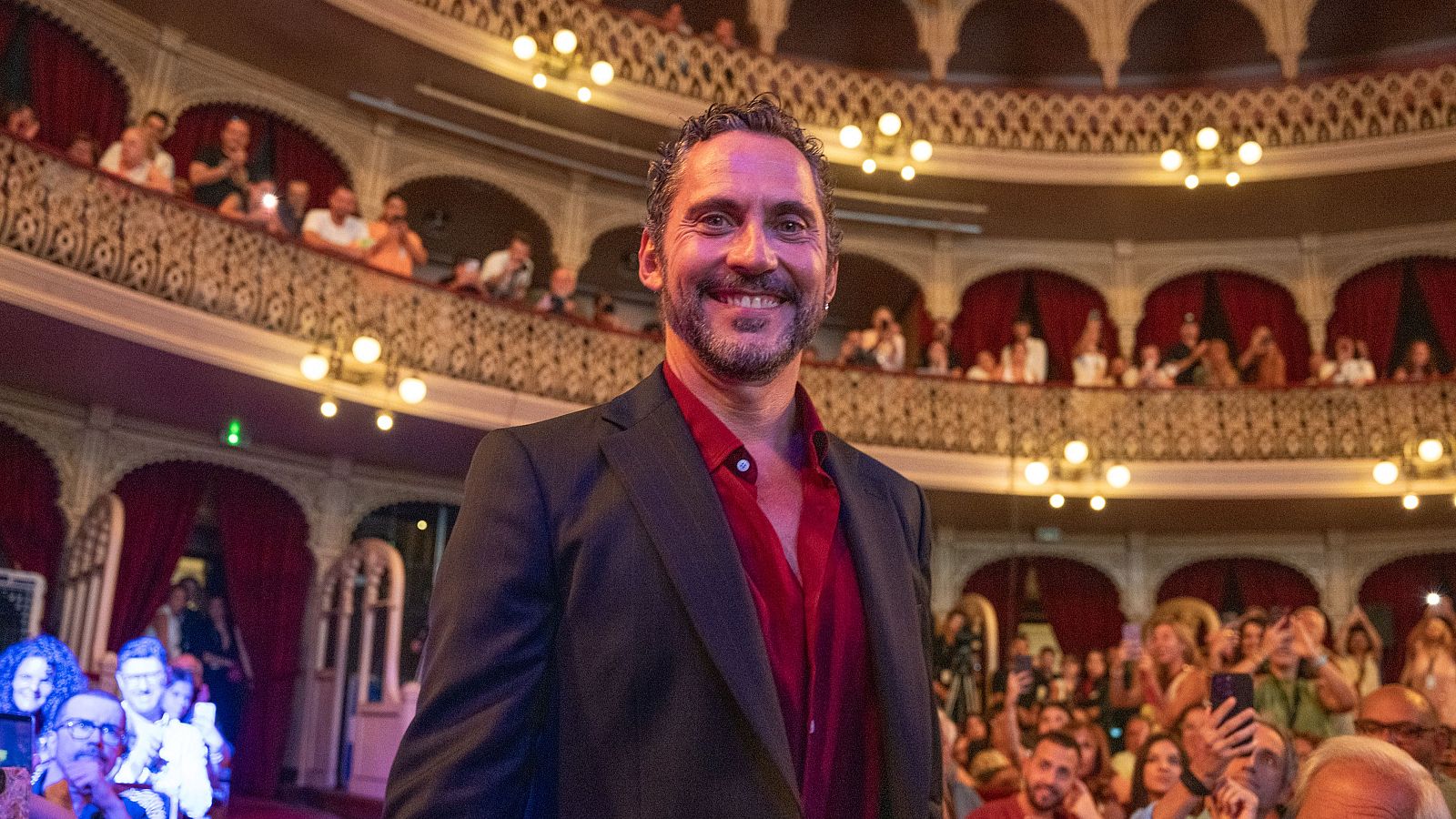 El actor Paco León, con americana oscura y camisa roja, sonríe al recibir un premio en el Gran Teatro Falla ante un público que aplaude.