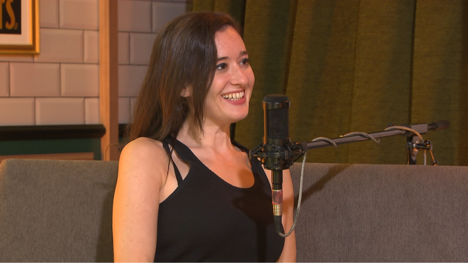 Mujer joven en estudio de radio, sonriendo ante un micrófono.  Cabello castaño, camiseta negra. Fondo con azulejos blancos y cortina oscura.