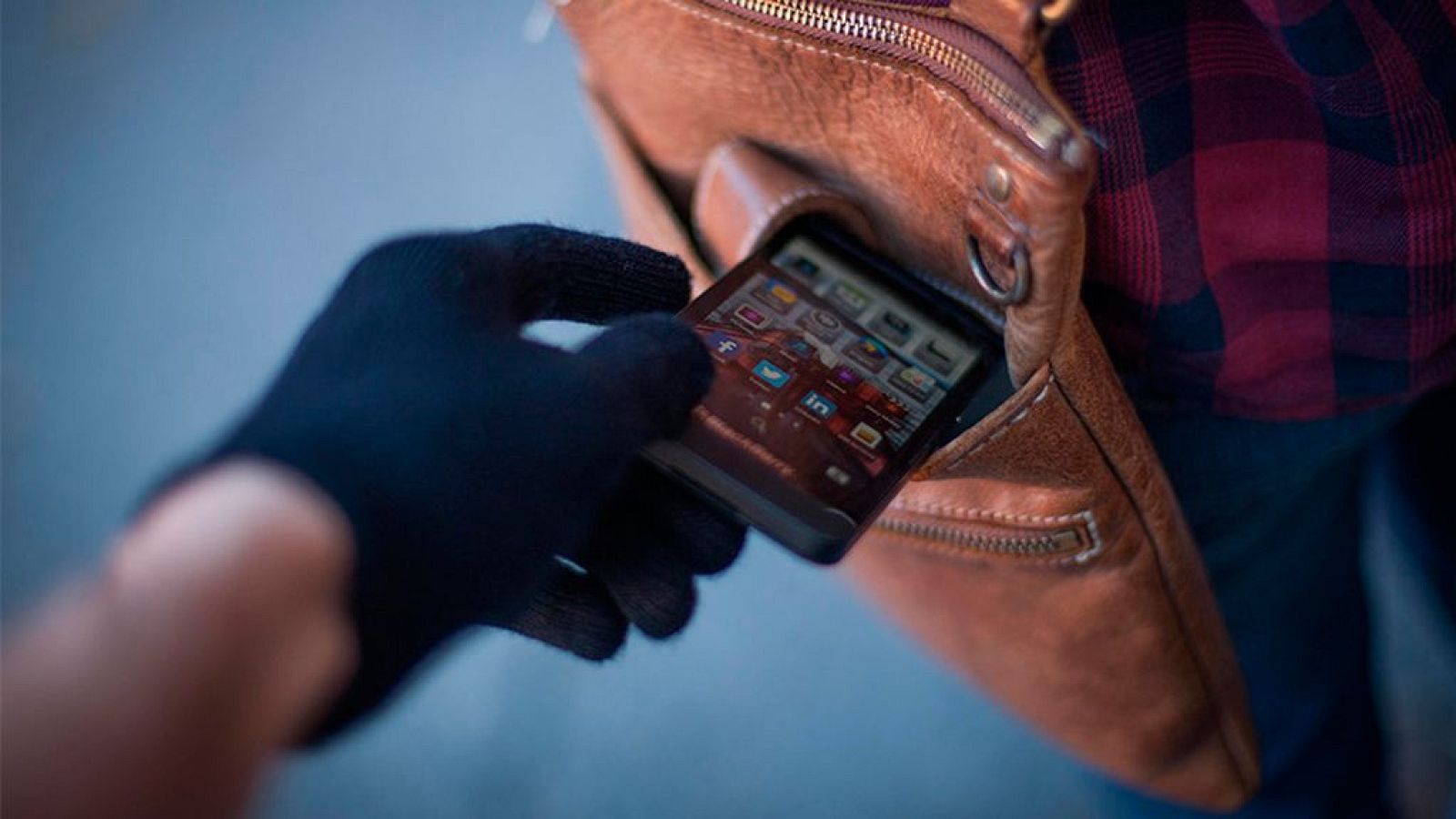 Robo de un smartphone de un bolso bandolera.  Mano enguantada extrae el teléfono con la pantalla encendida. Víctima viste camisa a cuadros.