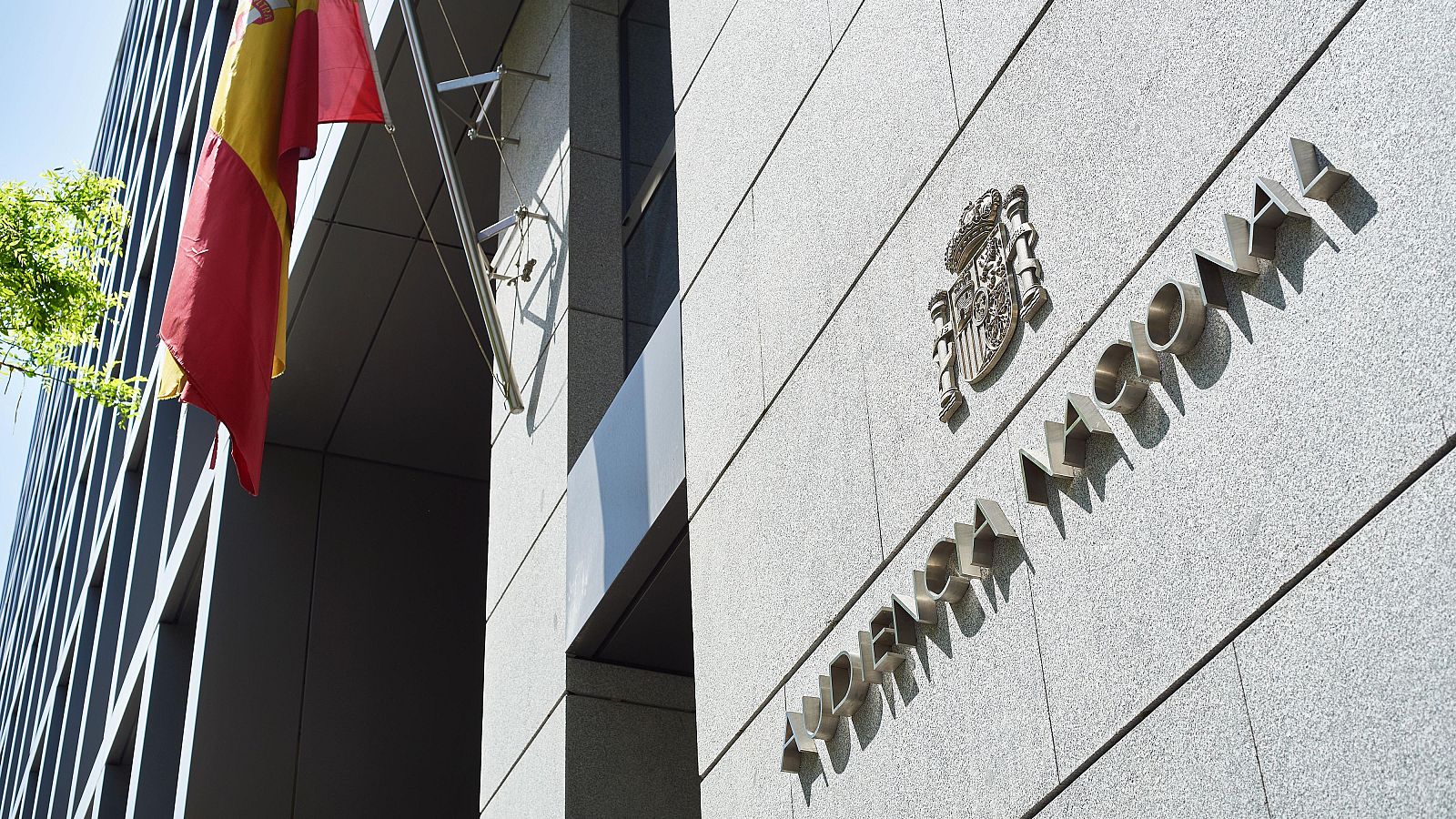 Fachada de la Audiencia Nacional: pared gris, letras plateadas con el nombre del edificio y bandera española.