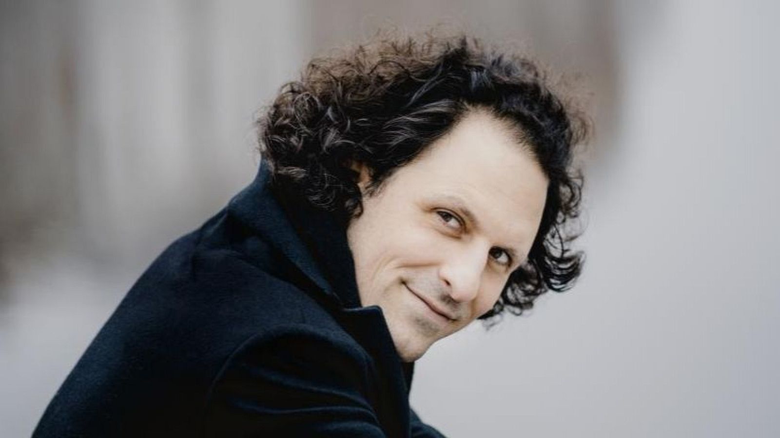 Retrato de un hombre con pelo oscuro y rizado, tez clara, chaqueta oscura y expresión serena. Mira a la cámara.